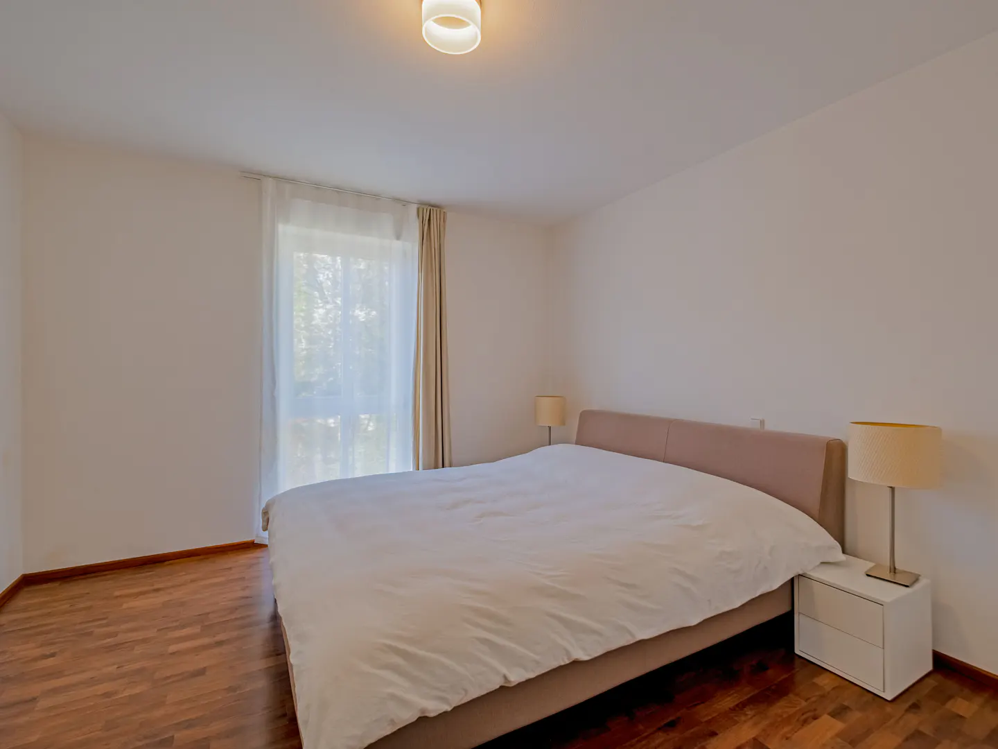 Bedroom with a king bed, white linens, and a white nightstand with a lamp. Hardwood floors and a window with sheer curtains.