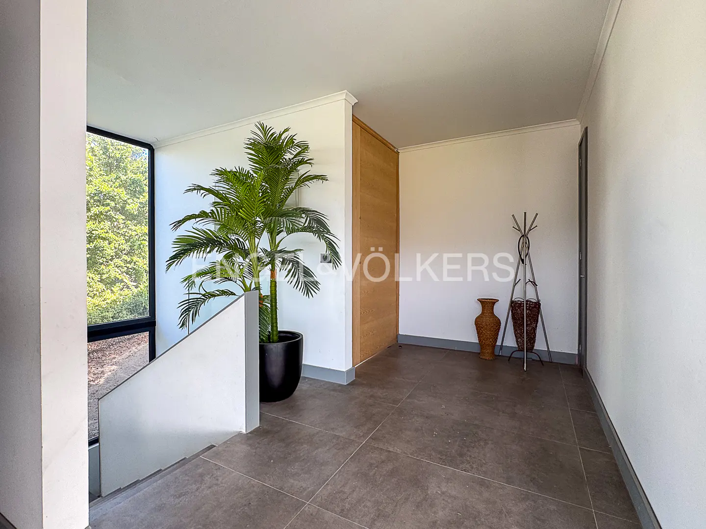 A modern home's entryway with a potted palm, coat rack, and vases on a gray tiled floor. A large window shows green trees outside.