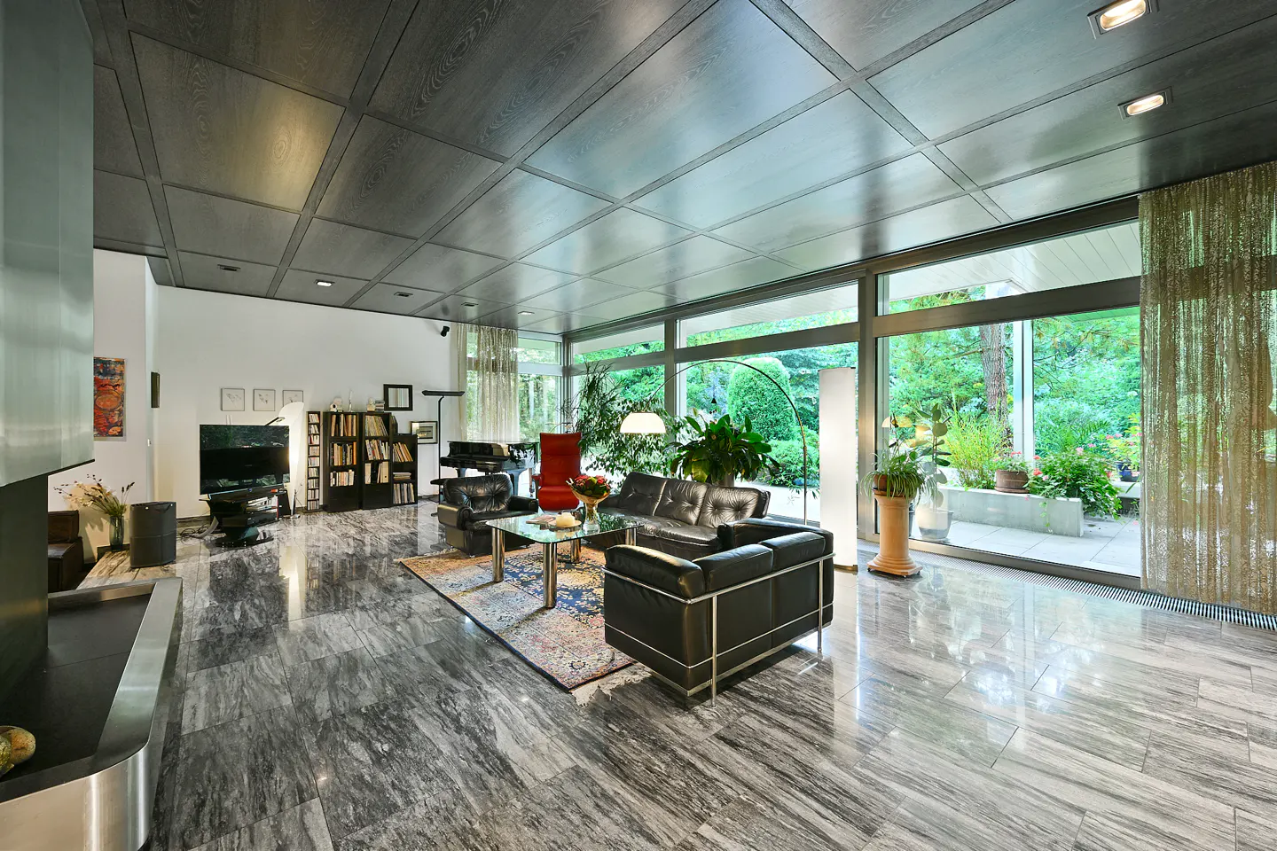 A modern living room with marble floors, a black leather sofa, and floor-to-ceiling windows overlooking a lush green garden.