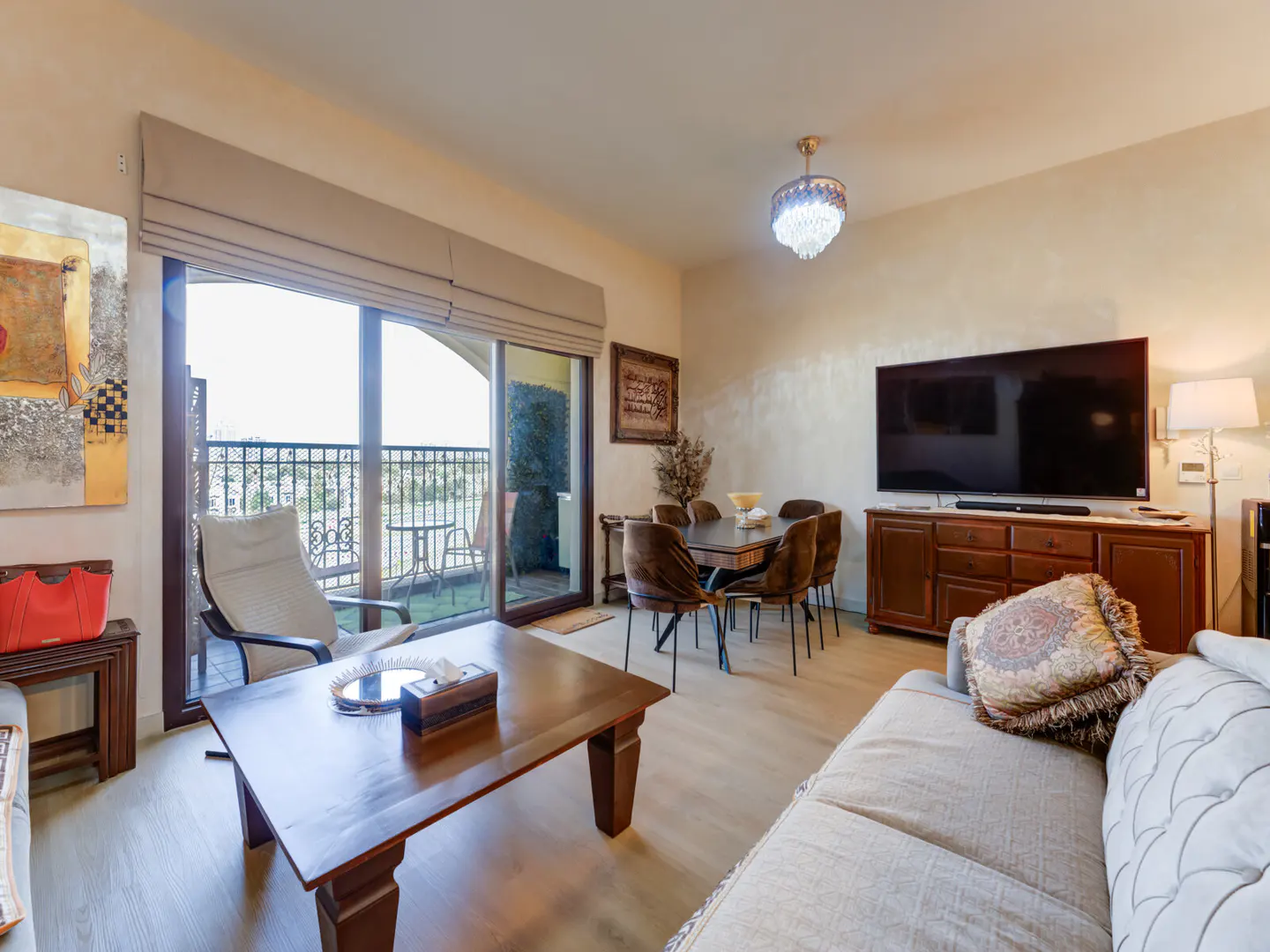 Living room with a beige sofa, wood table, dining set, and a balcony with a view. A large TV sits on a wooden cabinet.
