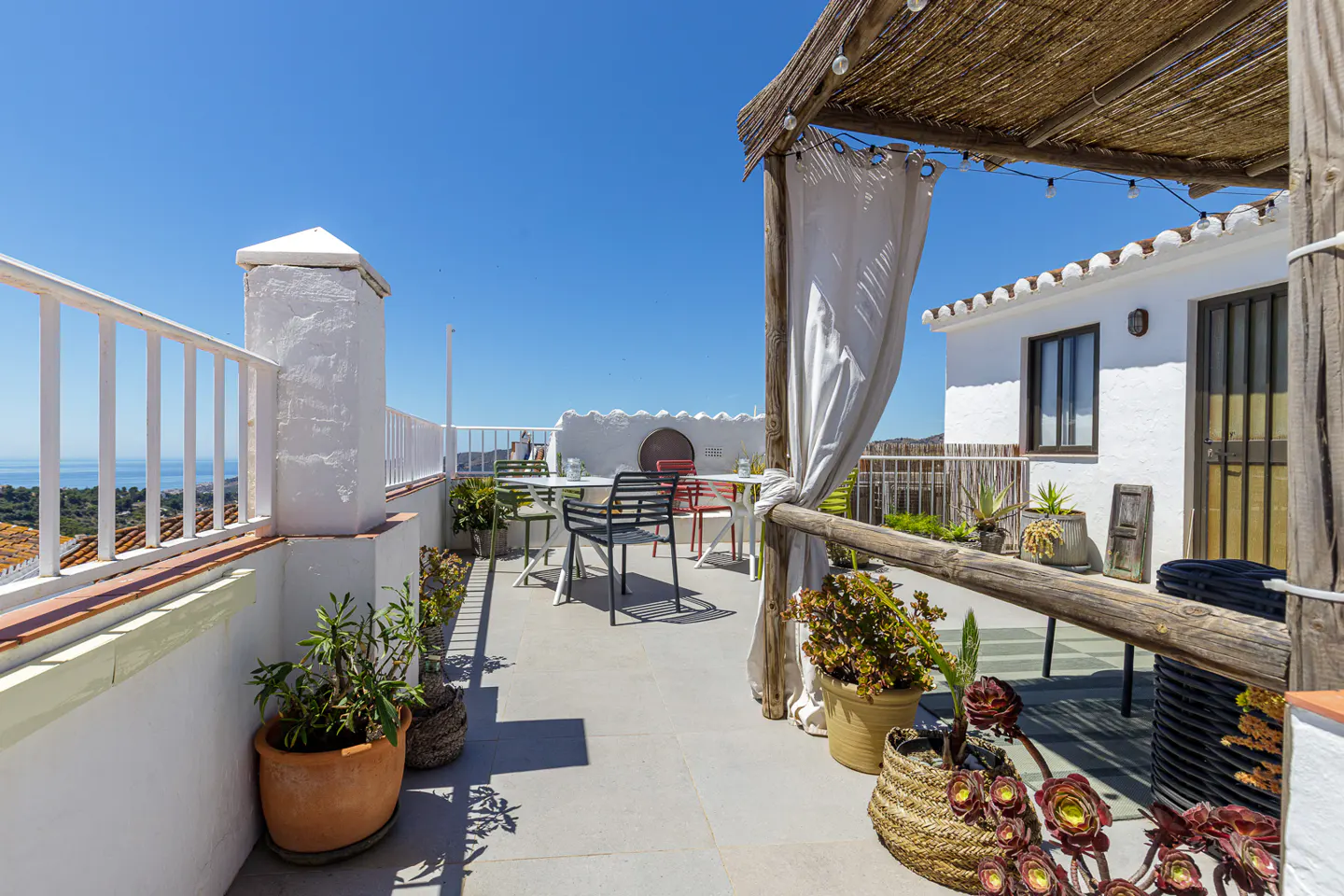 Sunny rooftop terrace with white walls, a wooden pergola, outdoor dining furniture, and potted plants overlooking the sea.