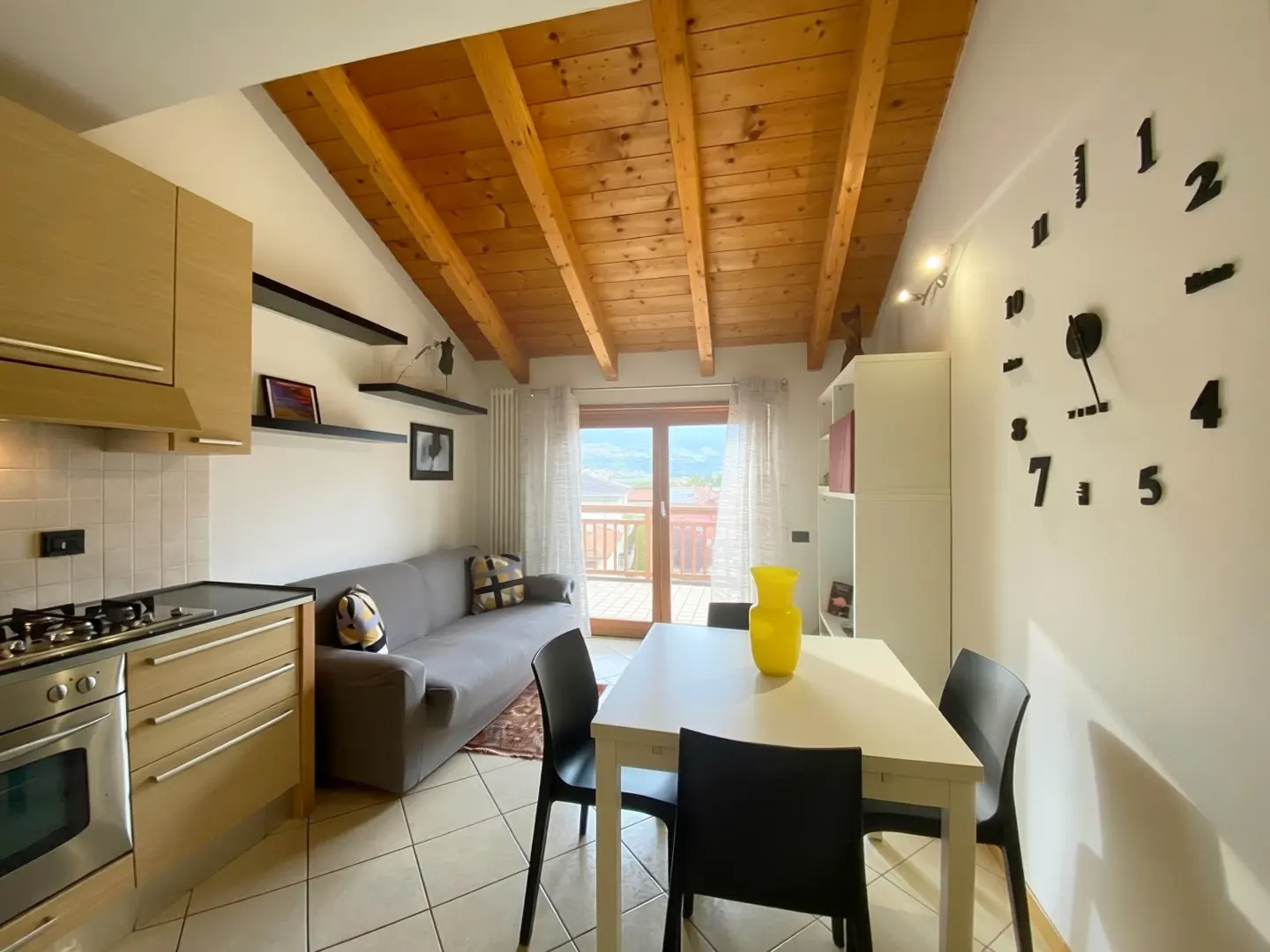 Cozy apartment interior with wood ceiling, kitchen, gray sofa, and dining table with black chairs. A large clock adorns the wall. Balcony view.