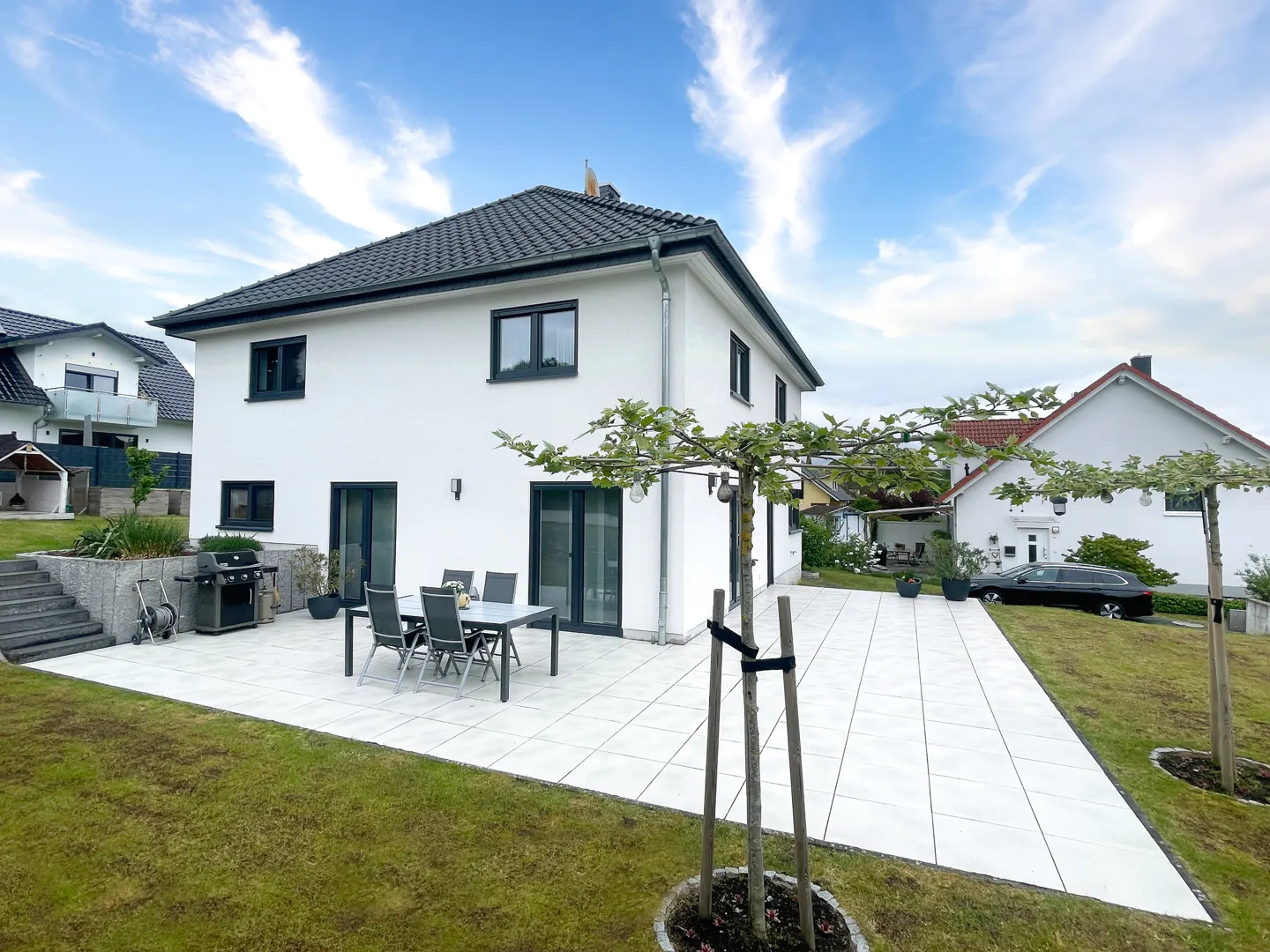 White two-story house with a tiled patio, outdoor dining set, grill, and young trees in a grassy backyard under blue sky.