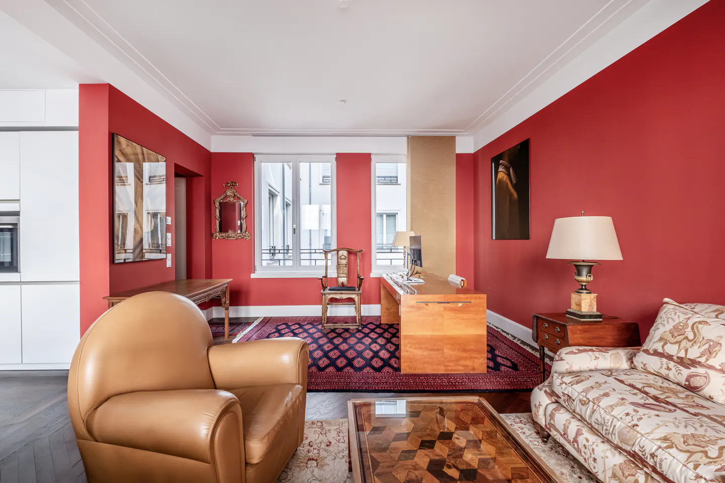 A living room with red walls, a wooden desk, a leather chair, and a patterned sofa. A window provides natural light.