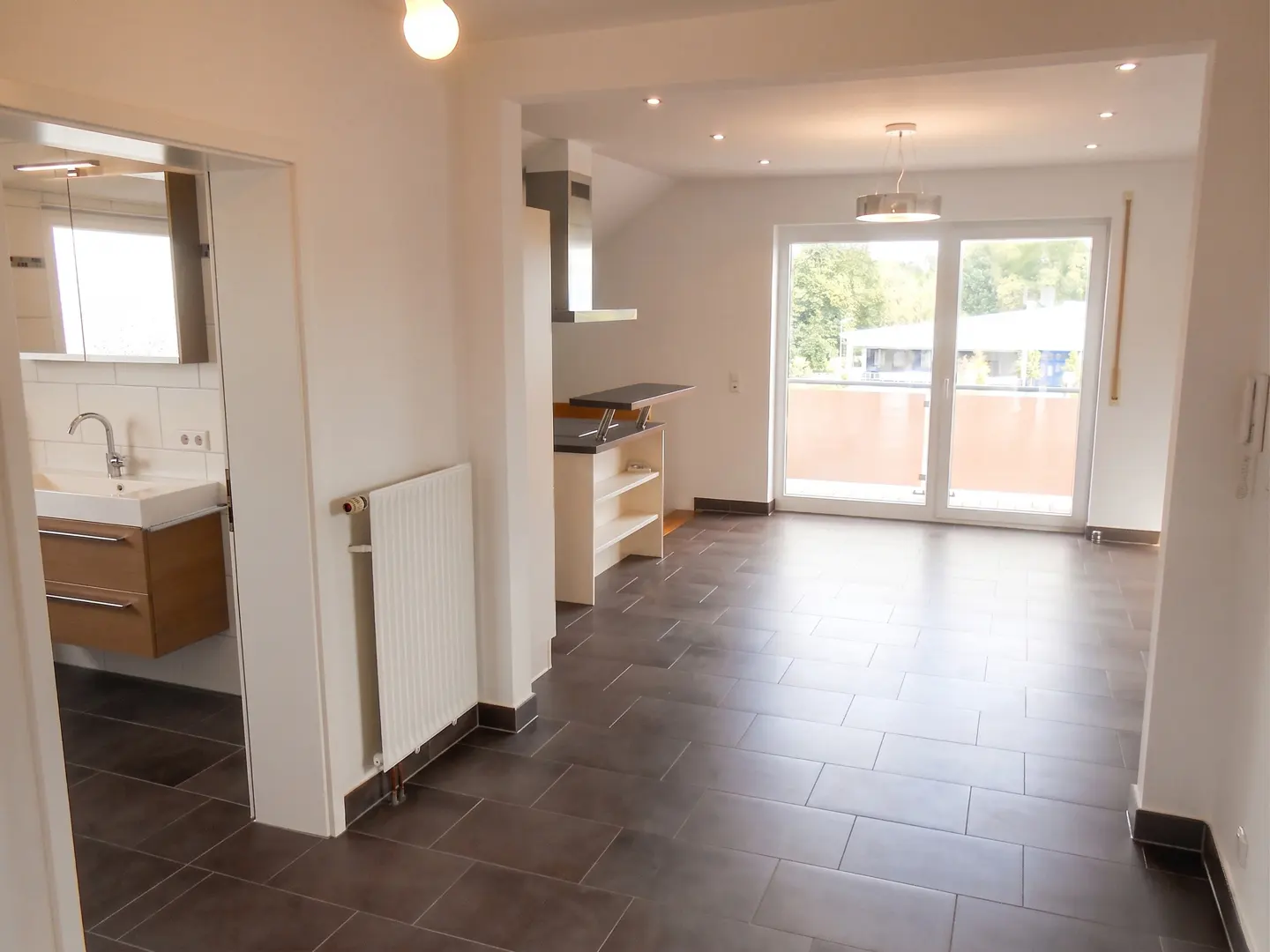 A bright, open-concept apartment with dark tile floors, a kitchen bar, and a balcony with a view. A bathroom is visible through a doorway.
