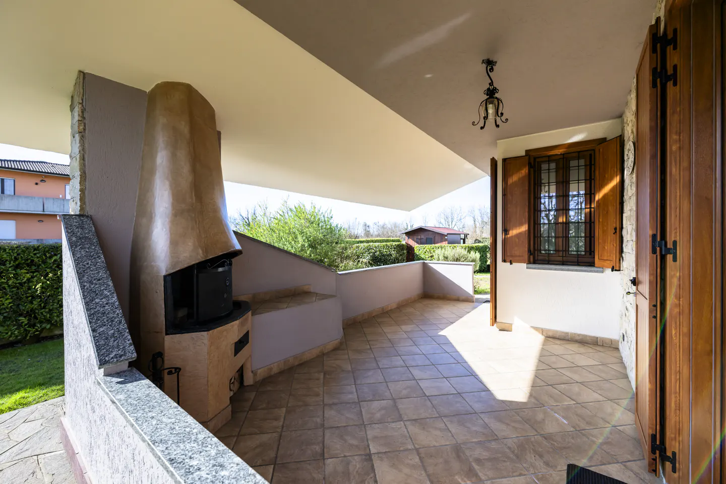 Covered patio with tile floor, stone fireplace, and wood shutters. Green lawn and trees are visible in the background.