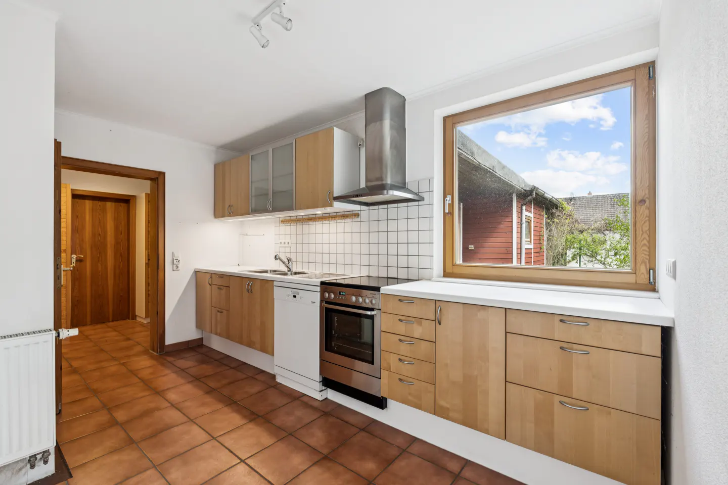 Bright kitchen with wood cabinets, stainless steel appliances, and terracotta tile floor. A window overlooks a red building and blue sky.