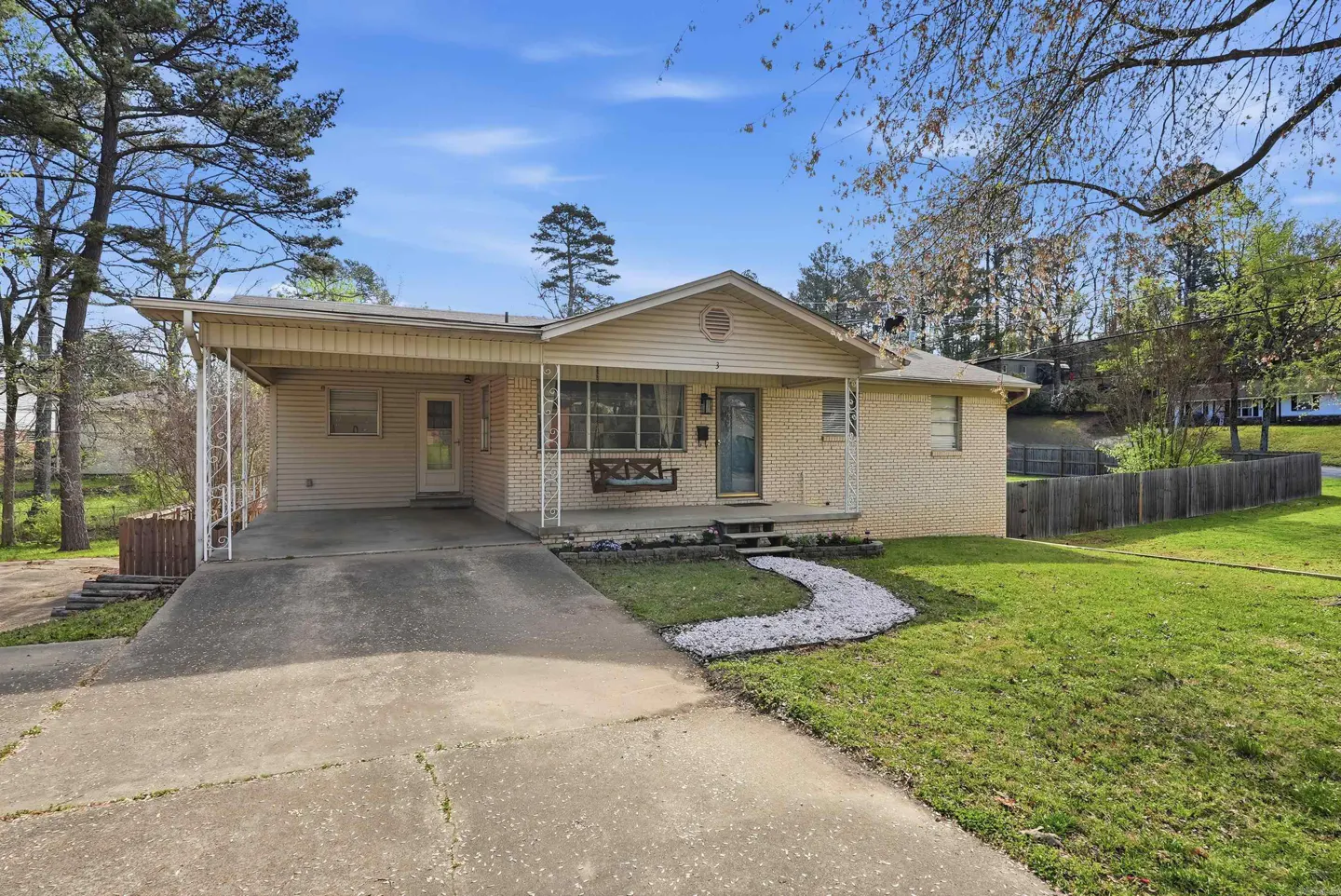 A single-story brick house with a carport, a small porch with a swing, and a concrete driveway. A white stone path leads to the lawn.