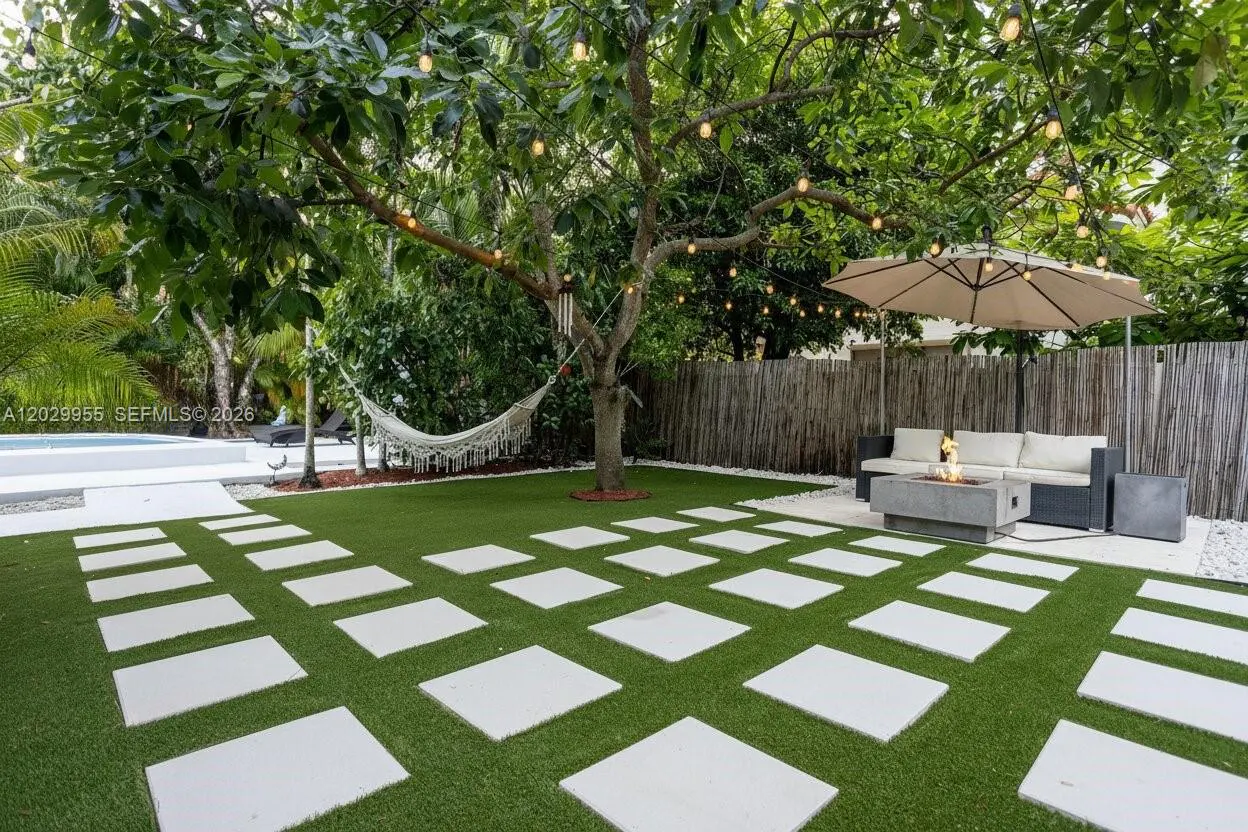 Lush backyard with artificial turf, square stone pavers, a hammock, fire pit, outdoor seating, and string lights.