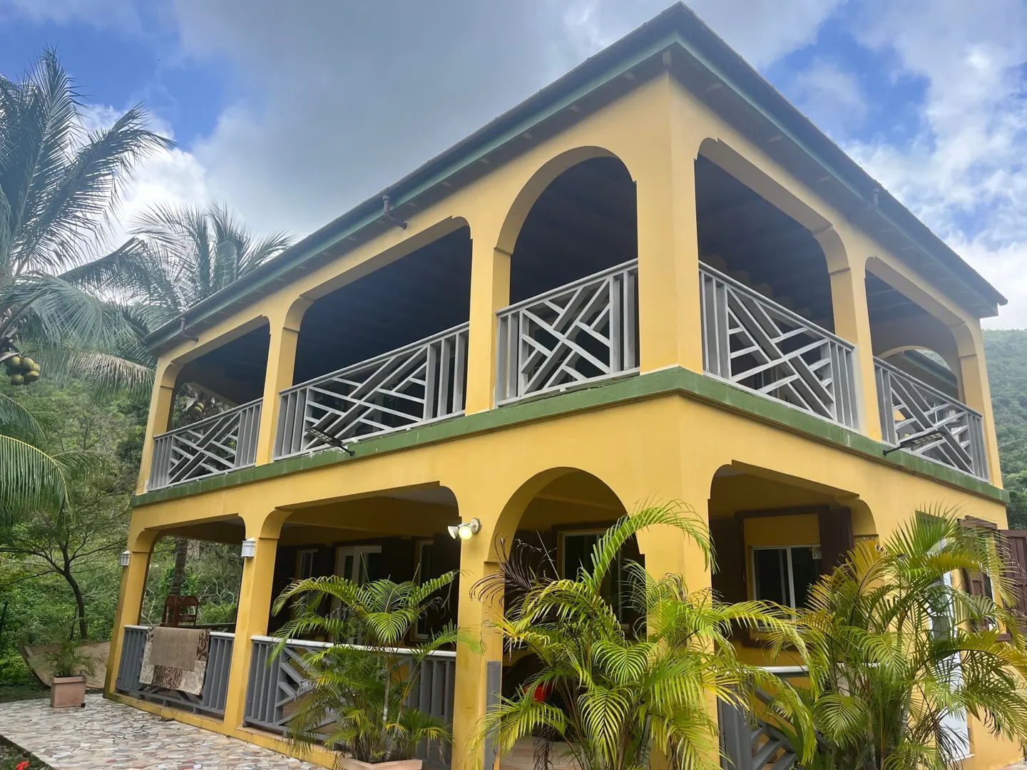 Two-story yellow house with arched balconies and white railings, surrounded by lush green tropical plants and trees.