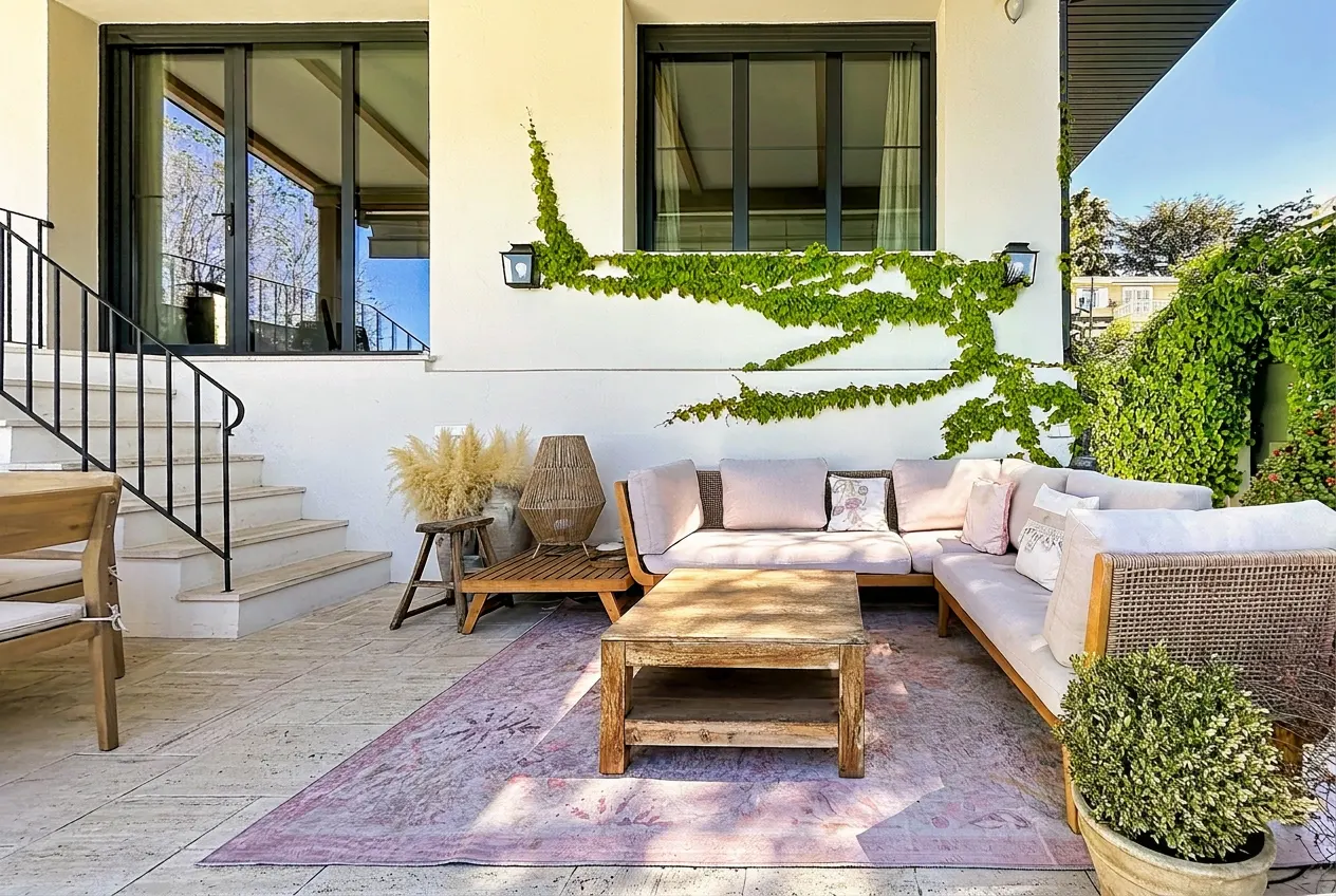 Outdoor patio with a light pink rug, wooden furniture, and climbing vines on a white wall.