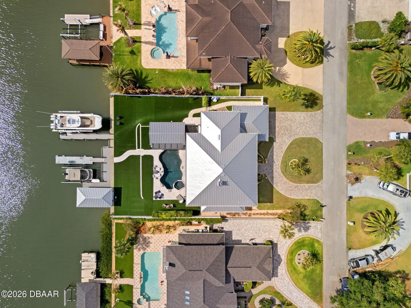 Aerial view of waterfront homes with pools, docks, and boats on a sunny day. Lush green lawns and palm trees surround the properties.