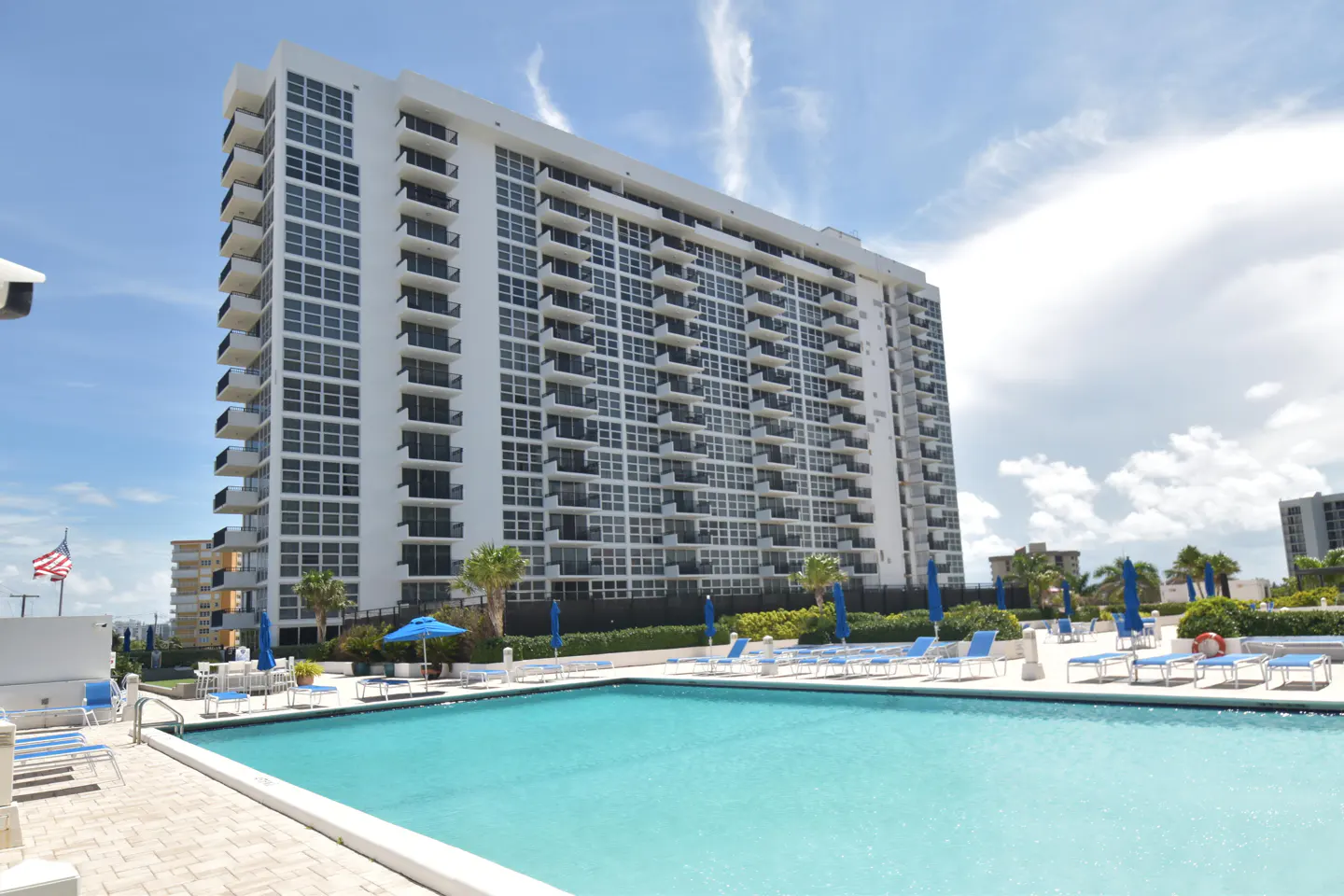 Exterior view of a tall white condo building with a pool, blue lounge chairs, and umbrellas on a sunny day.
