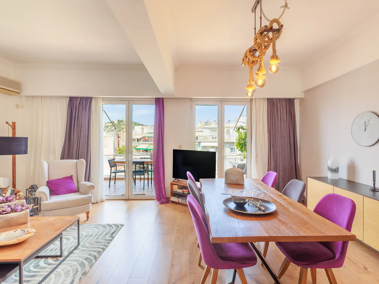 Bright living and dining area with wood floors, purple accents, and balcony access. A rope chandelier hangs above the dining table.