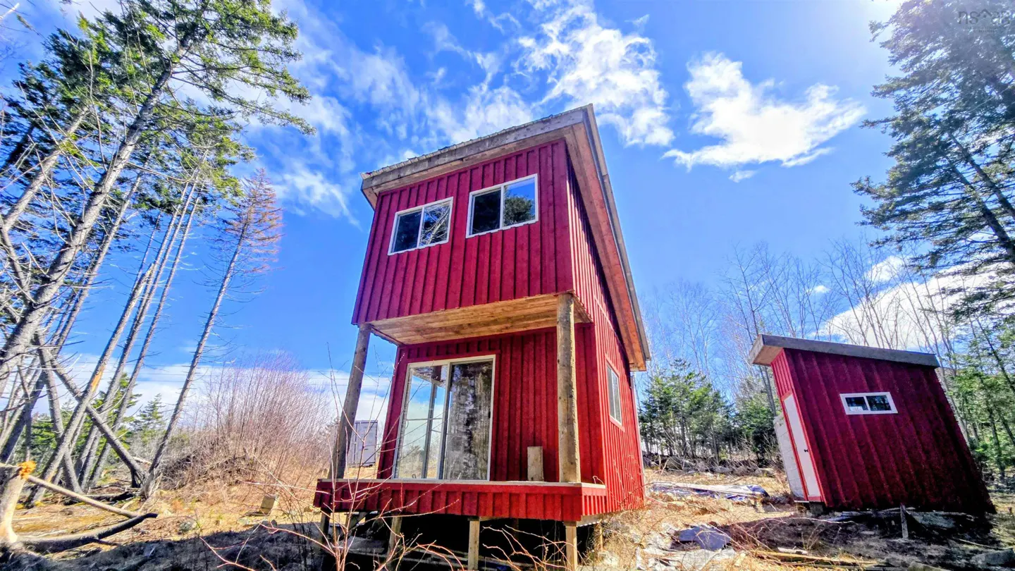 A two-story red cabin with a small porch, surrounded by trees under a blue sky with white clouds.