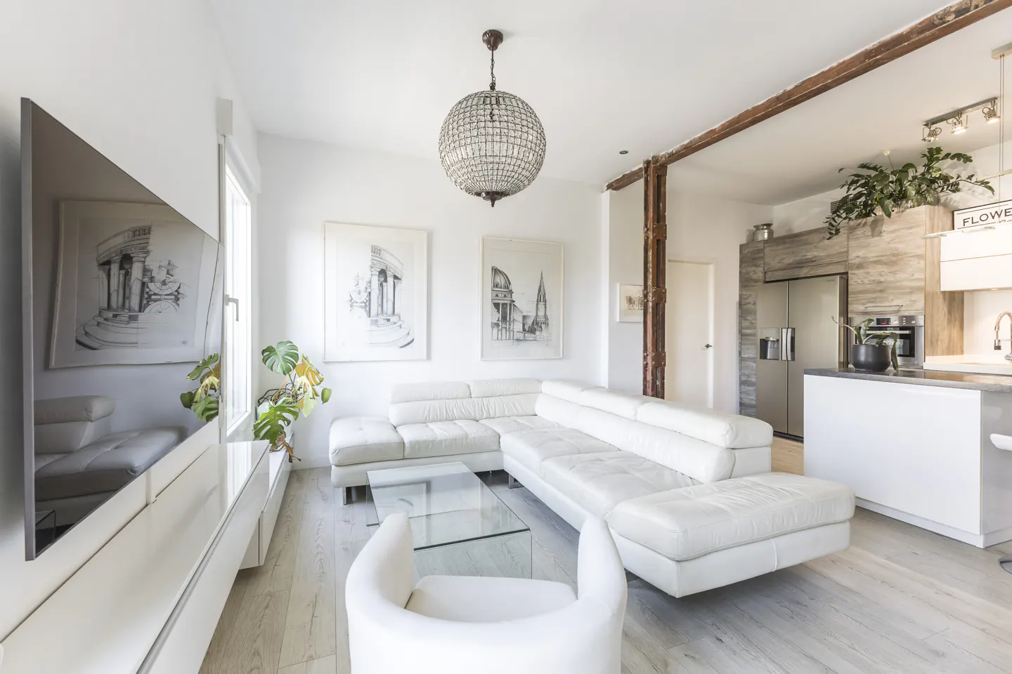 Modern living room with white sectional sofa, glass coffee table, crystal chandelier, and open-plan kitchen with wood accents.