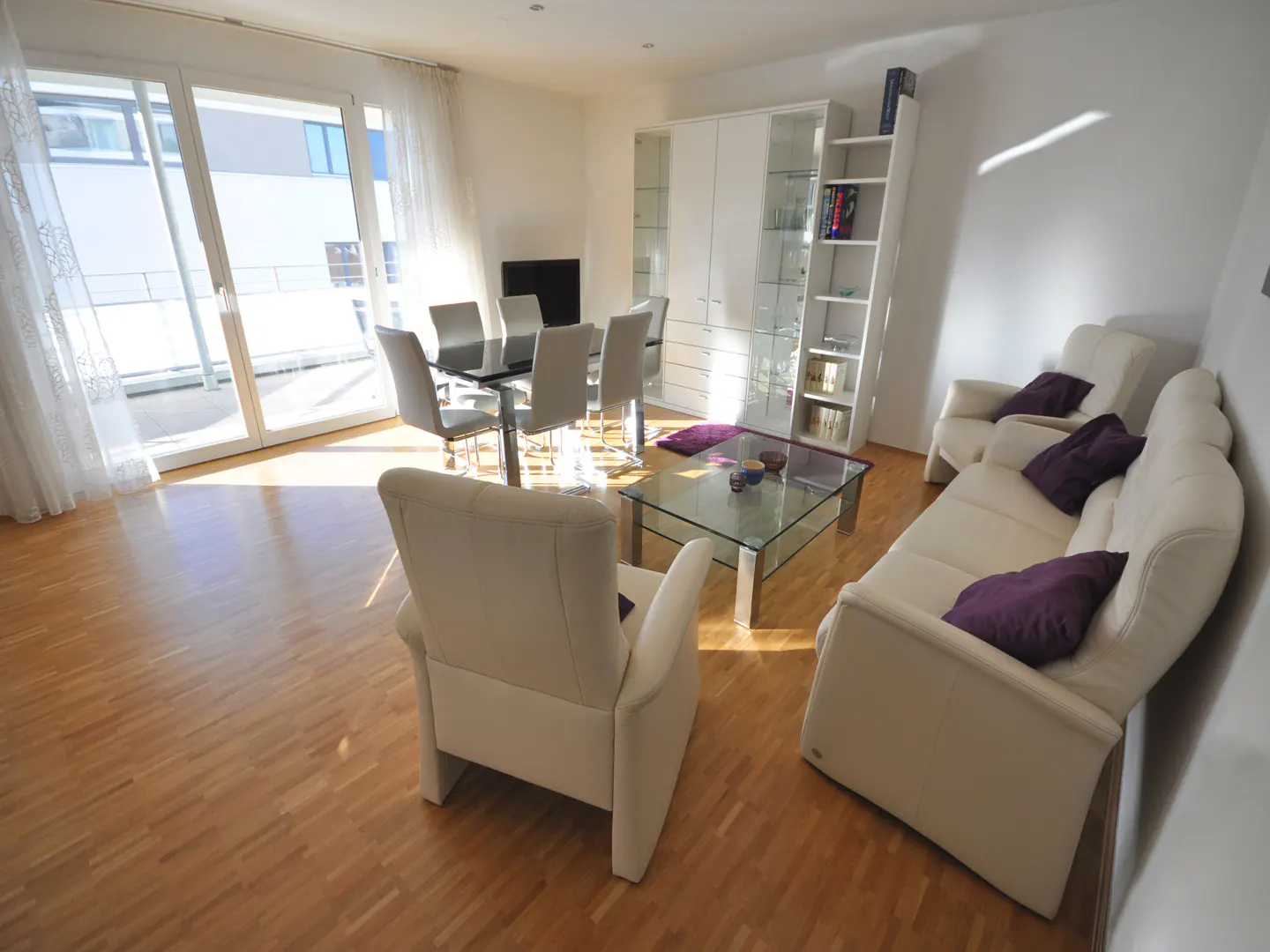 Bright living room with hardwood floors, white furniture, and a glass table. A dining table sits near large windows with sheer curtains.