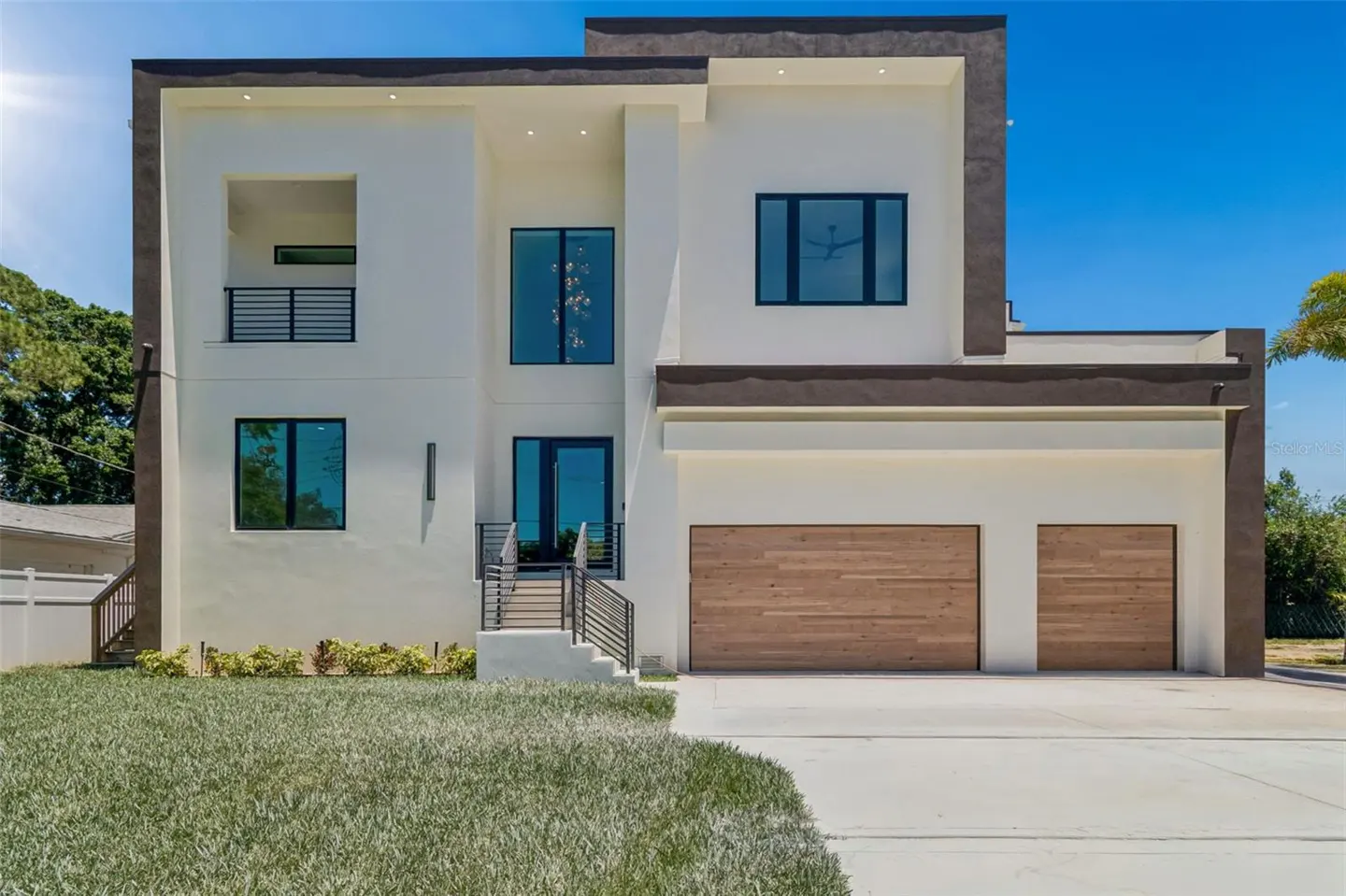 Two-story modern home with white stucco, dark trim, and wood garage doors. Lawn in front, blue sky above.