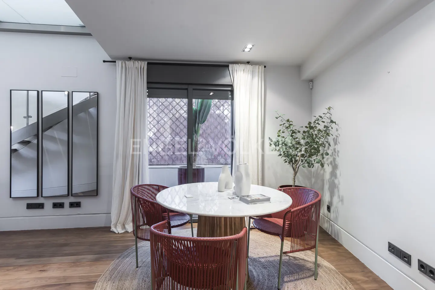 Bright dining area with a round marble table, four red woven chairs, and a jute rug. White walls, curtains, and a window with a view.