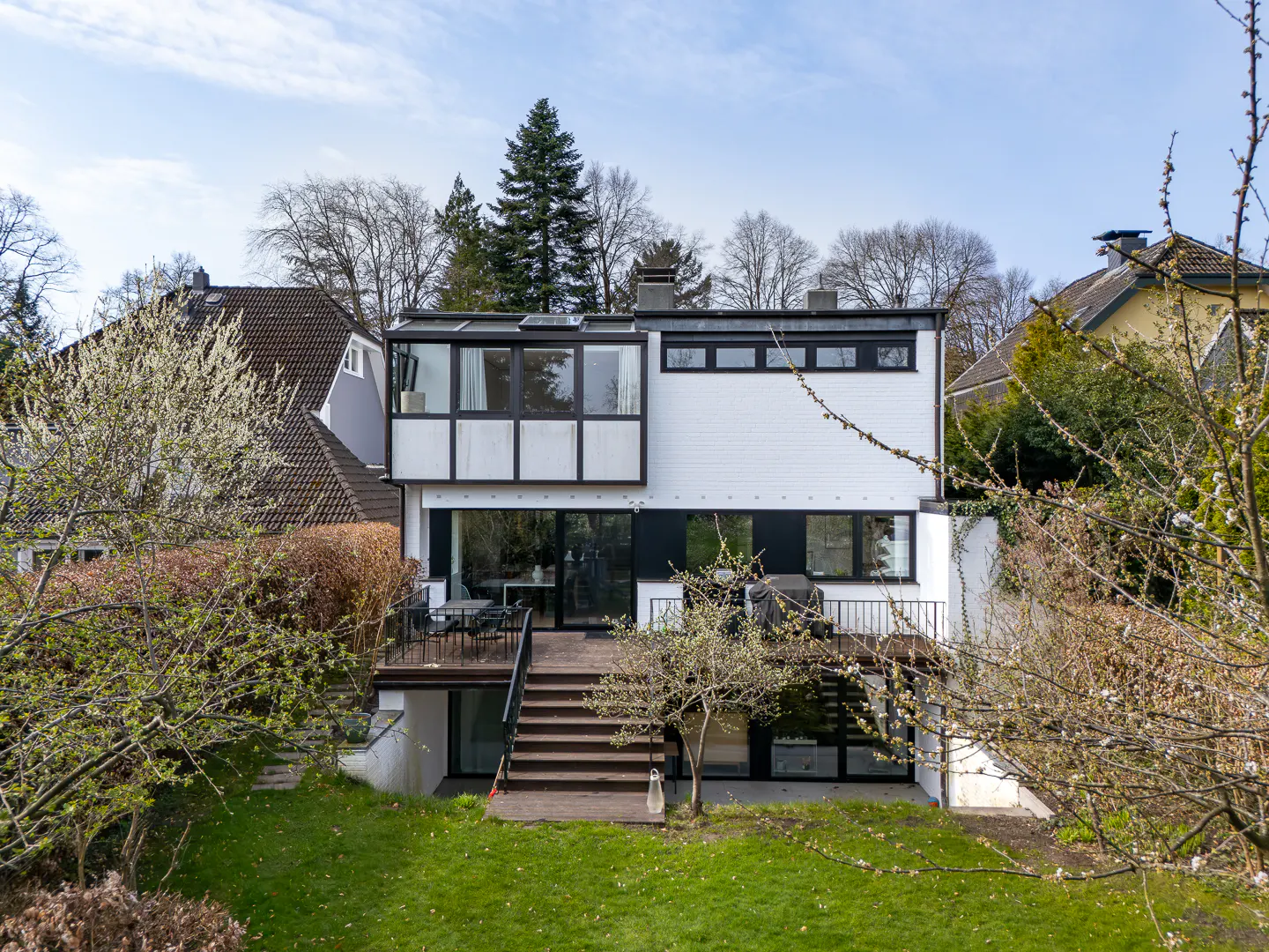 Modern white house with black trim, a wooden deck, and stairs leading to a green lawn. Trees surround the property.
