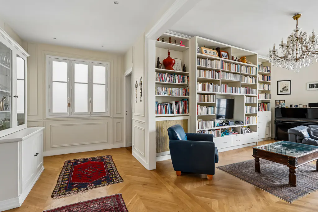 A bright living room with hardwood floors, a large bookcase, a blue armchair, and a crystal chandelier.