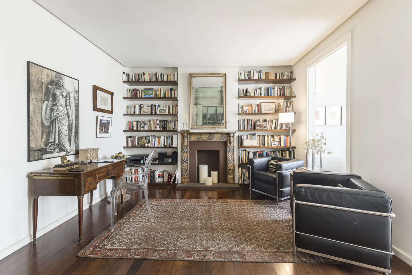 A home office with a wooden desk, two black leather armchairs, a patterned rug, and floor-to-ceiling bookshelves.