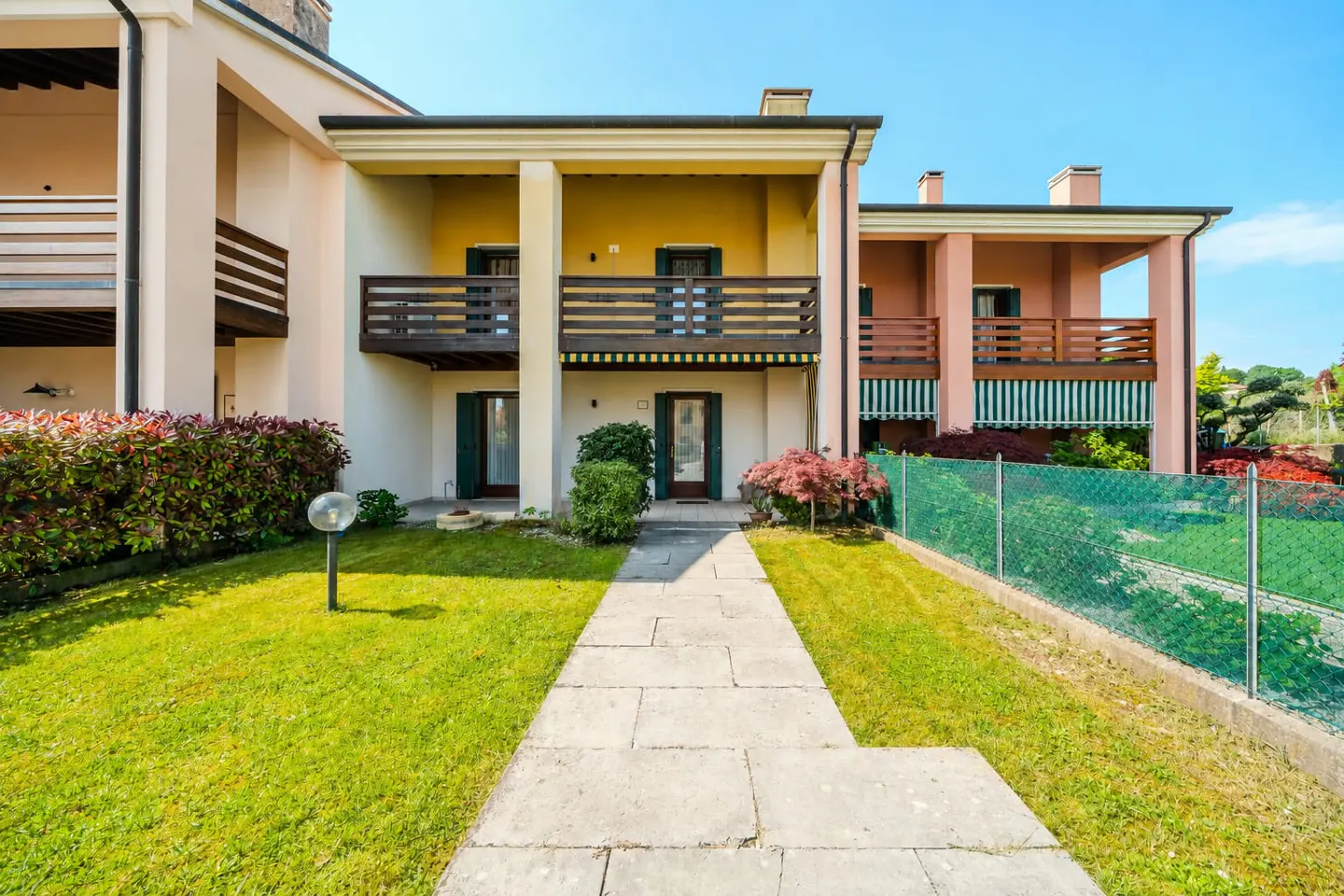 Exterior view of a two-story townhouse with balconies, a stone walkway, and a green lawn under a clear blue sky.