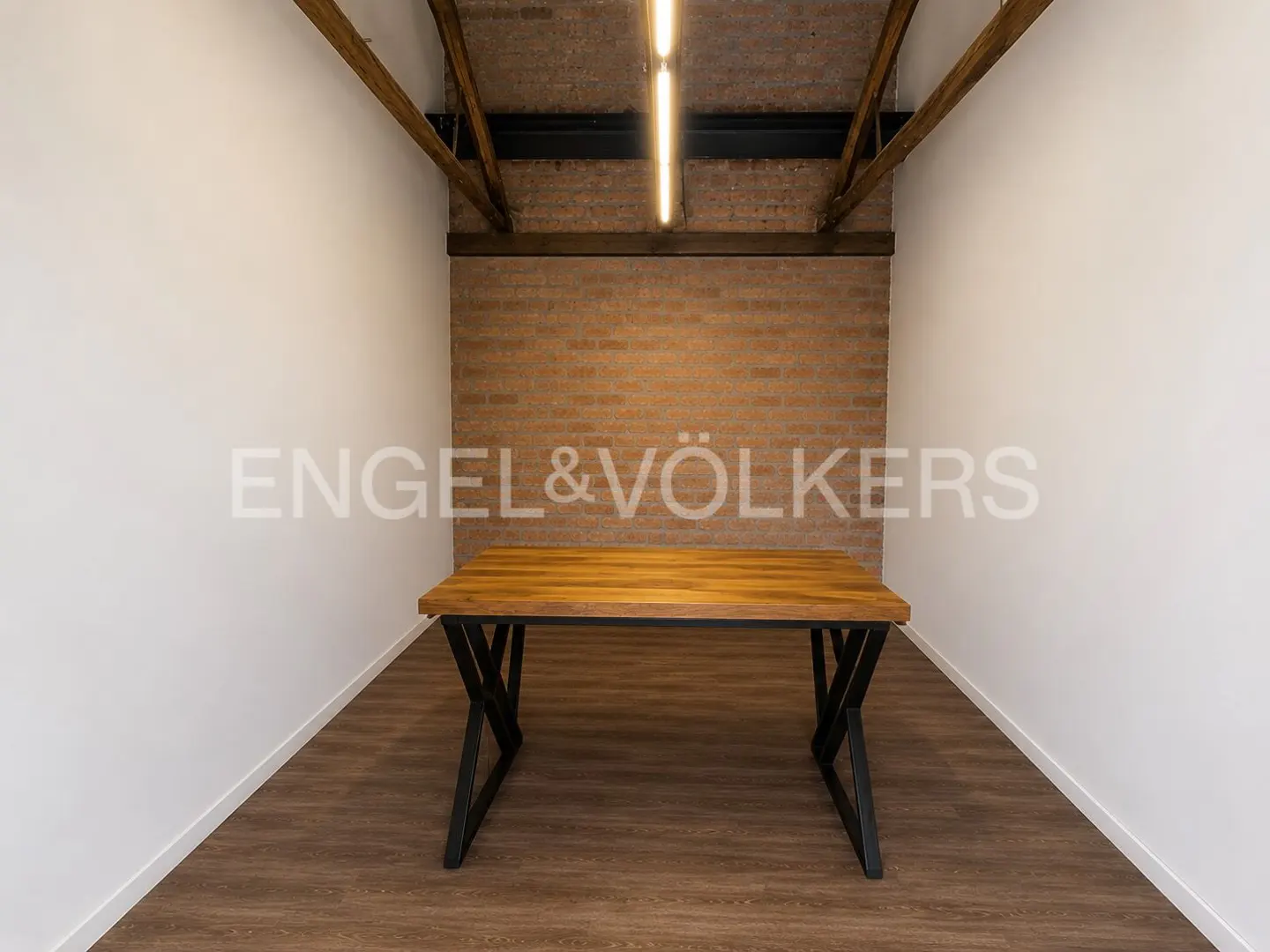 A wooden table with black metal legs sits in a room with white walls, a brick back wall, and exposed wooden ceiling beams.