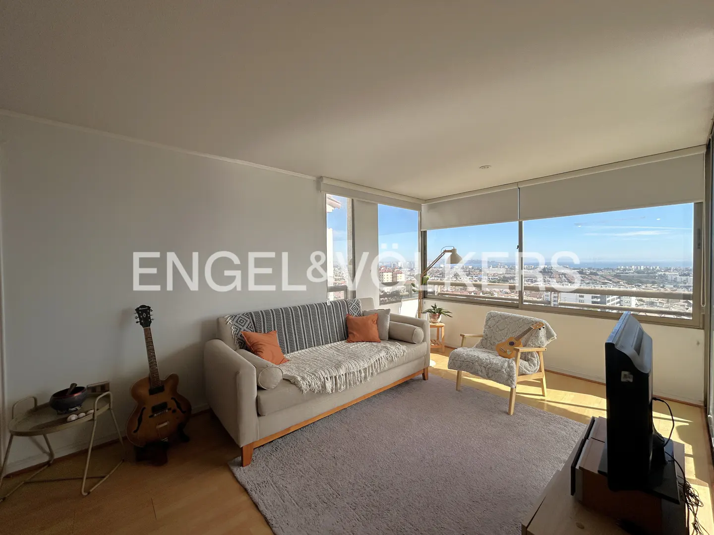Living room with a beige sofa, patterned chair, guitar, and a large window overlooking a city.