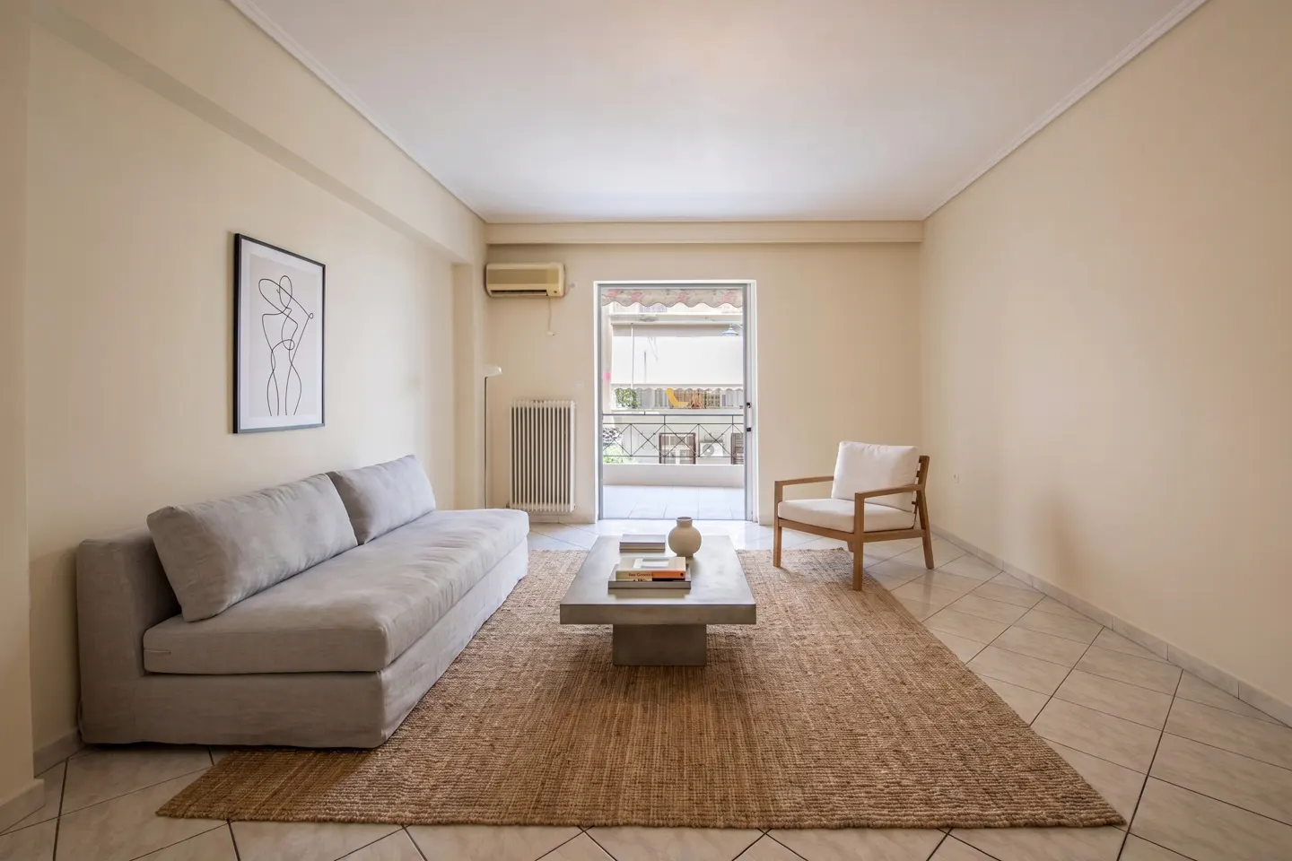 Minimalist living room with a grey sofa, wooden armchair, coffee table on a jute rug, and a balcony door.