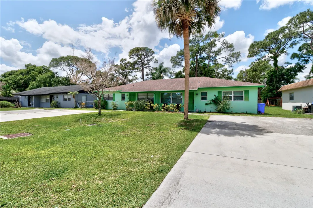 A light green single-story house with a brown roof, green shutters, and a palm tree in the front yard.