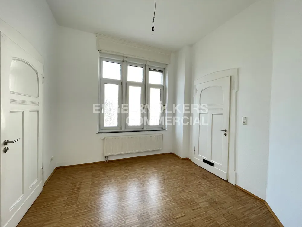 Bright, empty room with parquet floors, white walls, and two white doors. A window with white trim sits above a radiator. Engel & Volkers logo is superimposed on the window.