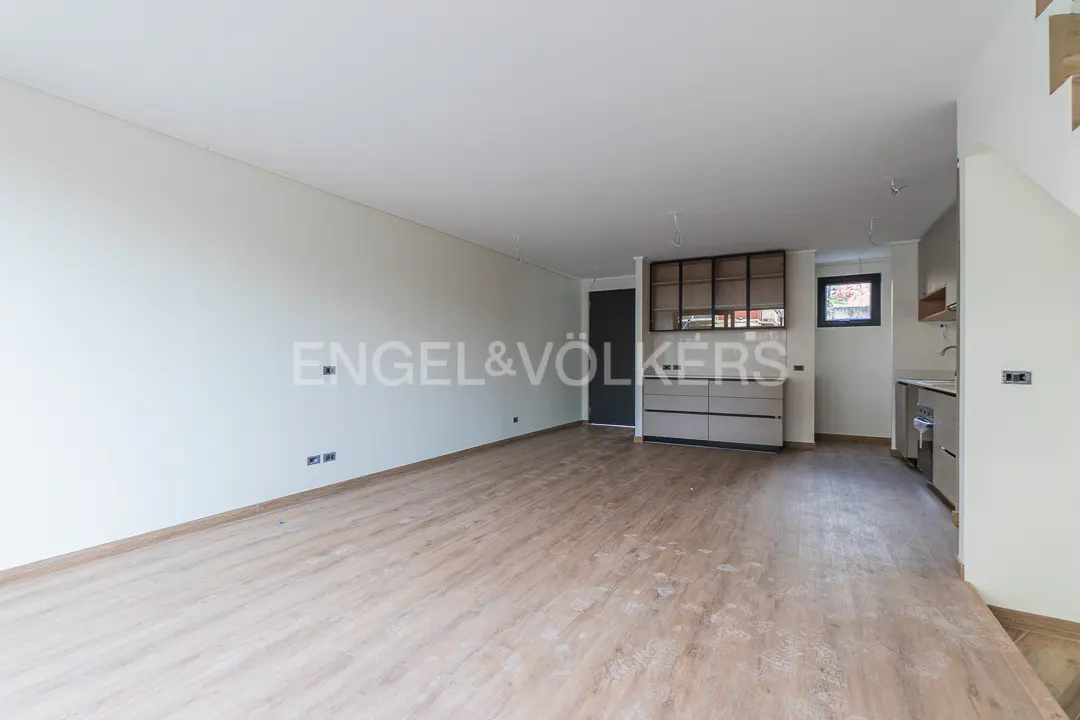 Open-plan living area with light wood floors, white walls, and a modern kitchen with grey cabinets and glass shelving.