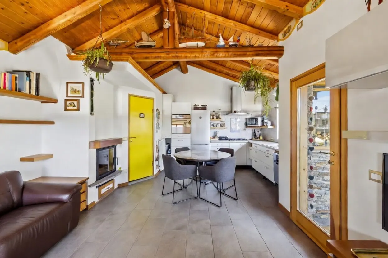 Open-concept living space with a wood-beamed ceiling, white walls, and a yellow door. A kitchen, dining area, and fireplace are visible.