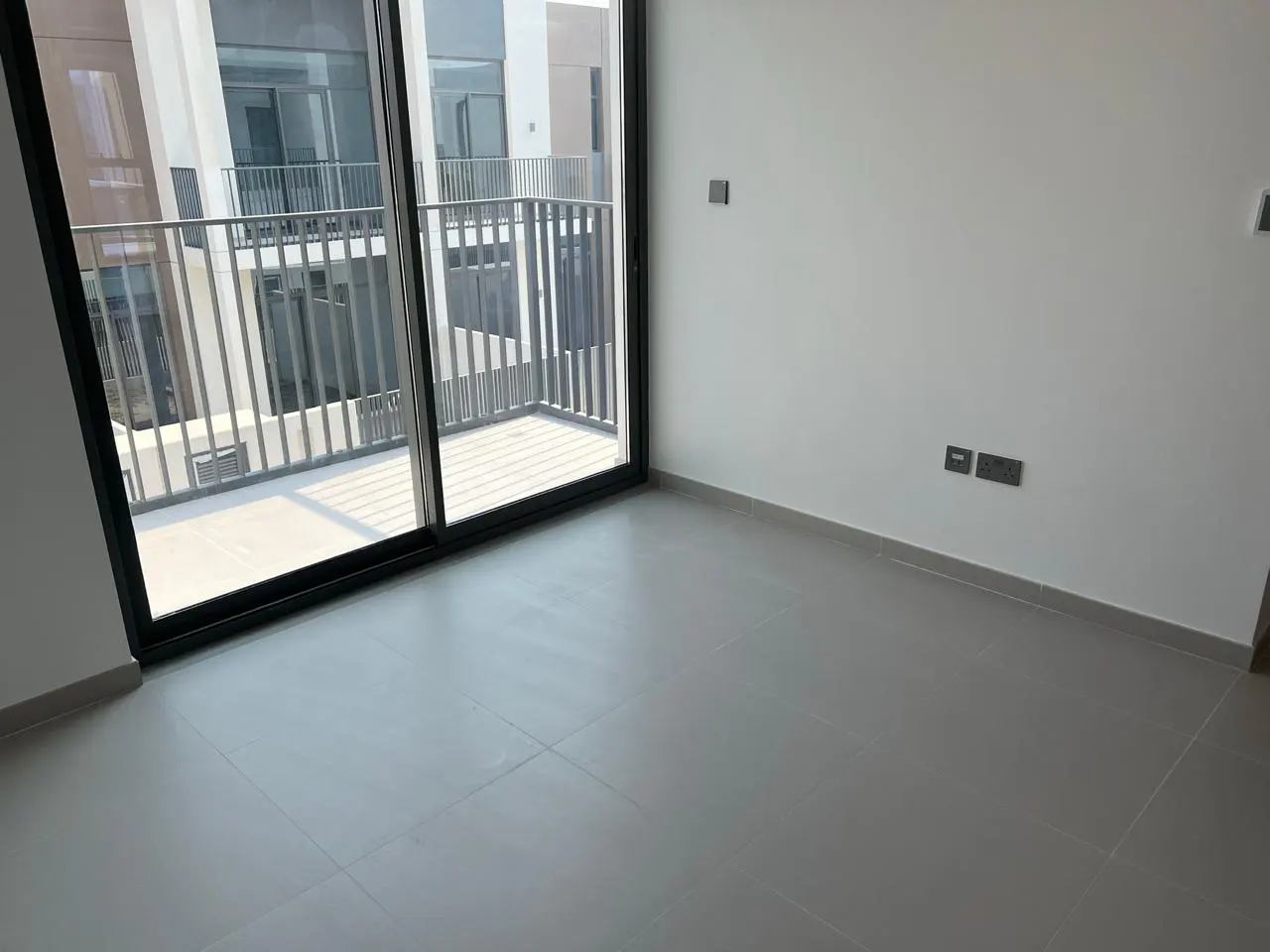 An empty room with gray tile floors and white walls. A sliding glass door leads to a balcony with metal railings.