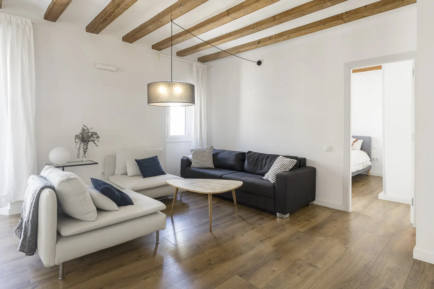 Bright living room with wood floors, white walls, and exposed beams. A black sofa and white chaise lounge face a light wood coffee table.