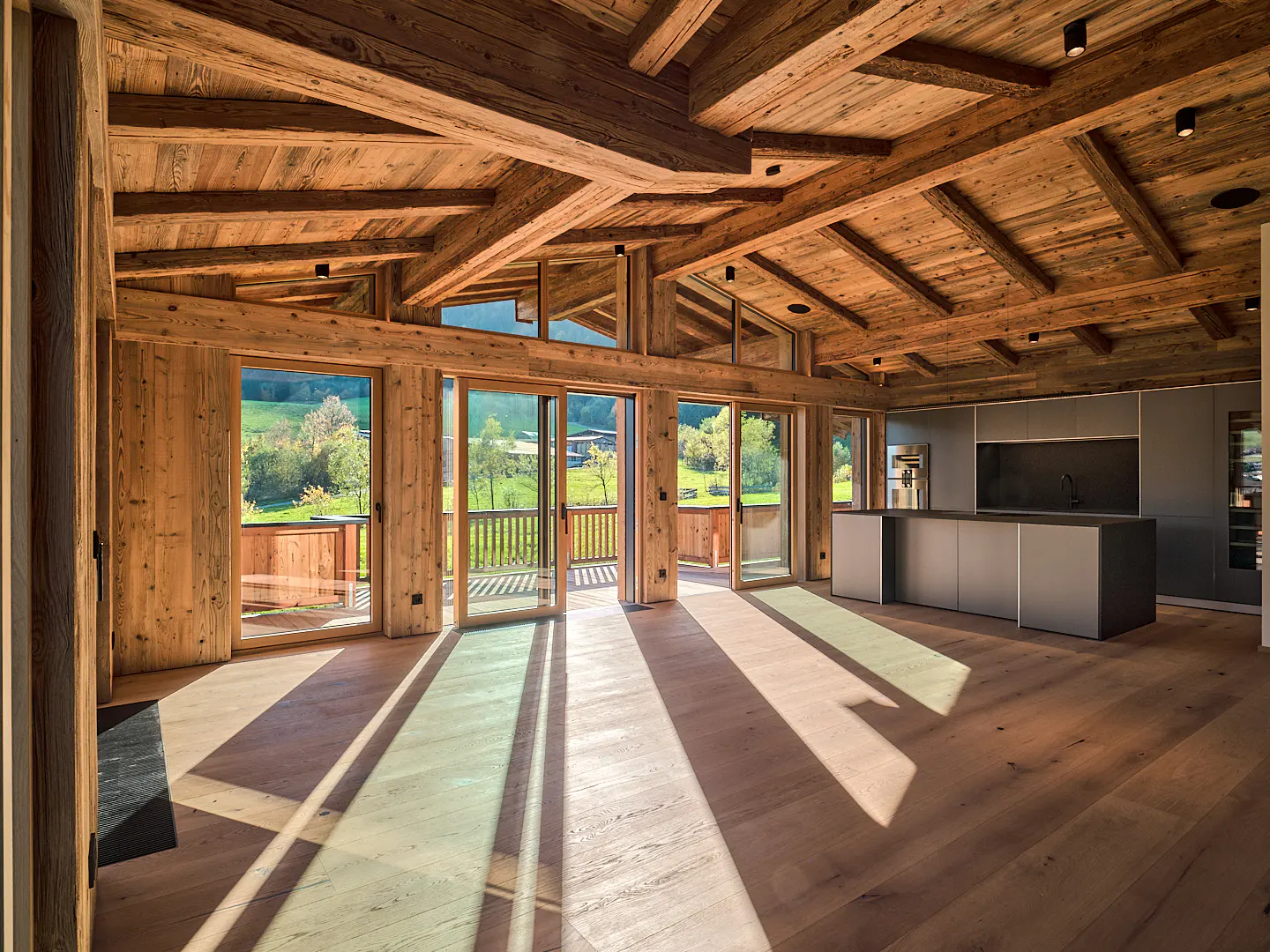 A bright, open-concept living space with wood floors, beams, and a modern kitchen. Sliding glass doors lead to a balcony with a green view.