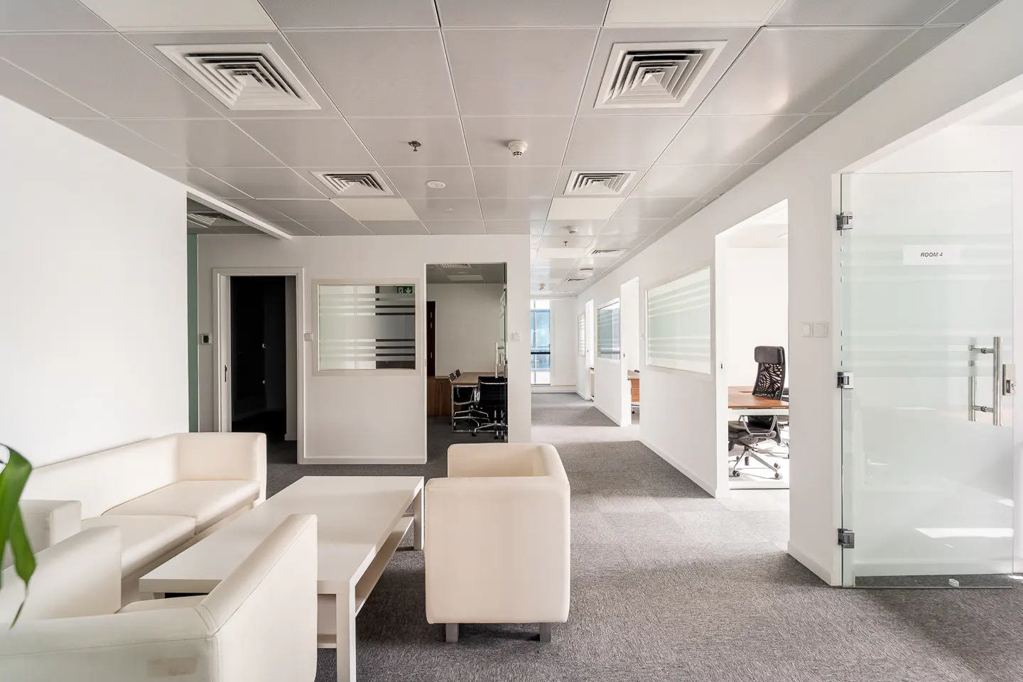 A bright office space with white walls, gray carpet, and a white sofa and table in the foreground. Glass-walled offices line the hallway.