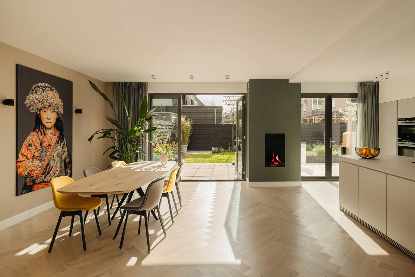 Bright, modern dining room with a long wooden table, yellow and gray chairs, and a large portrait. Open doors lead to a green backyard.