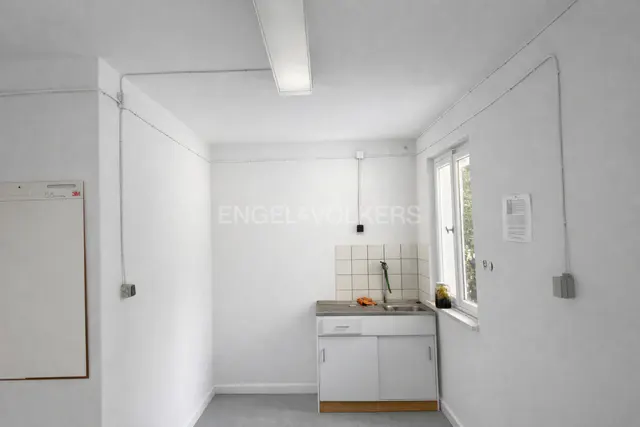Bright, empty room with white walls, grey floor, and a small white kitchen sink unit under a window. Engel & Völkers logo on the wall.