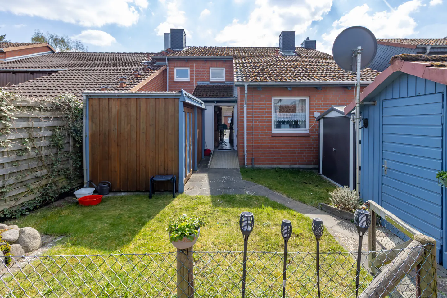 A backyard with a brick house, sheds, a fence, and solar lights on a sunny day.