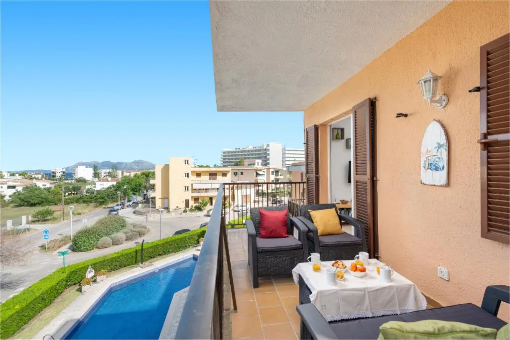 Balcony view with breakfast table, chairs, and pool. Peach wall with brown shutters, surfboard decor. Cityscape background under a clear blue sky.