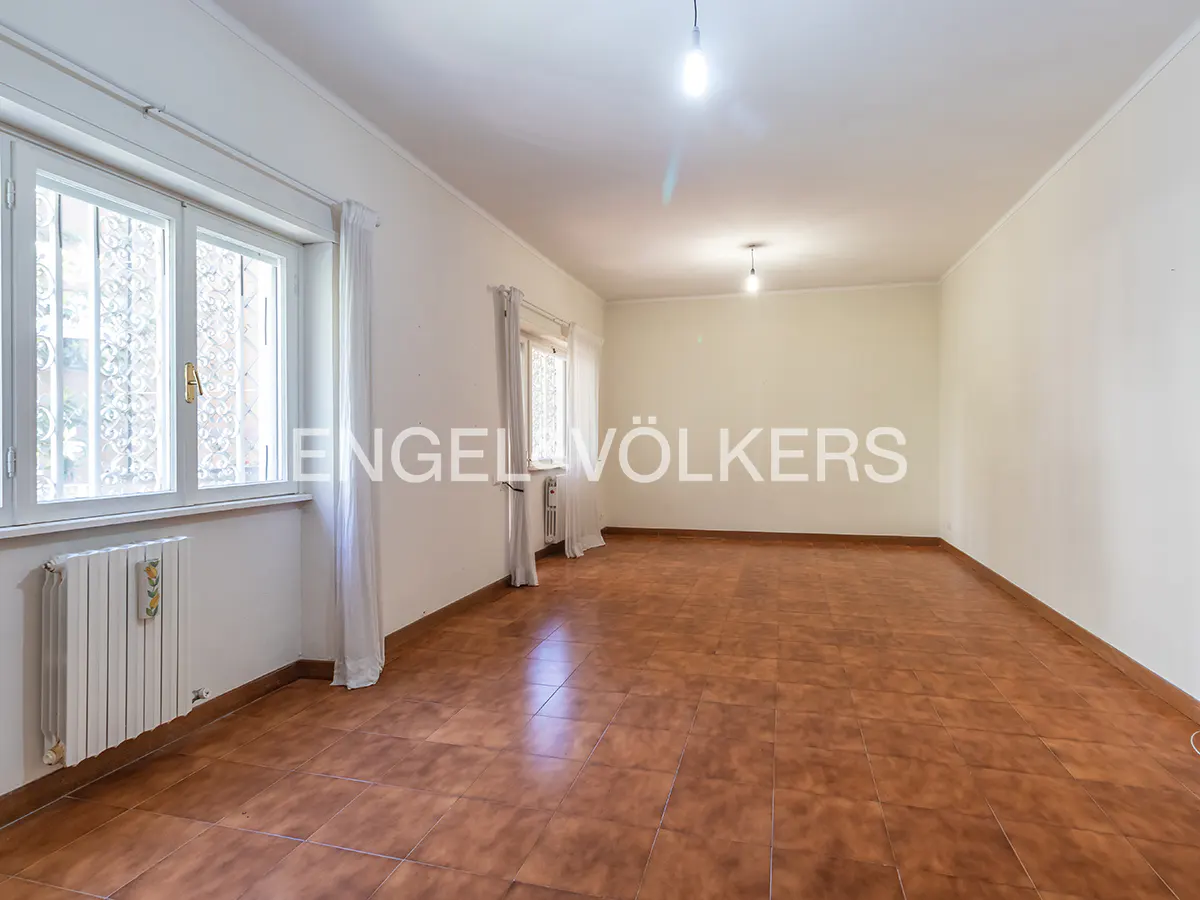 Empty room with white walls, brown tiled floors, two windows with white curtains, and a white radiator.