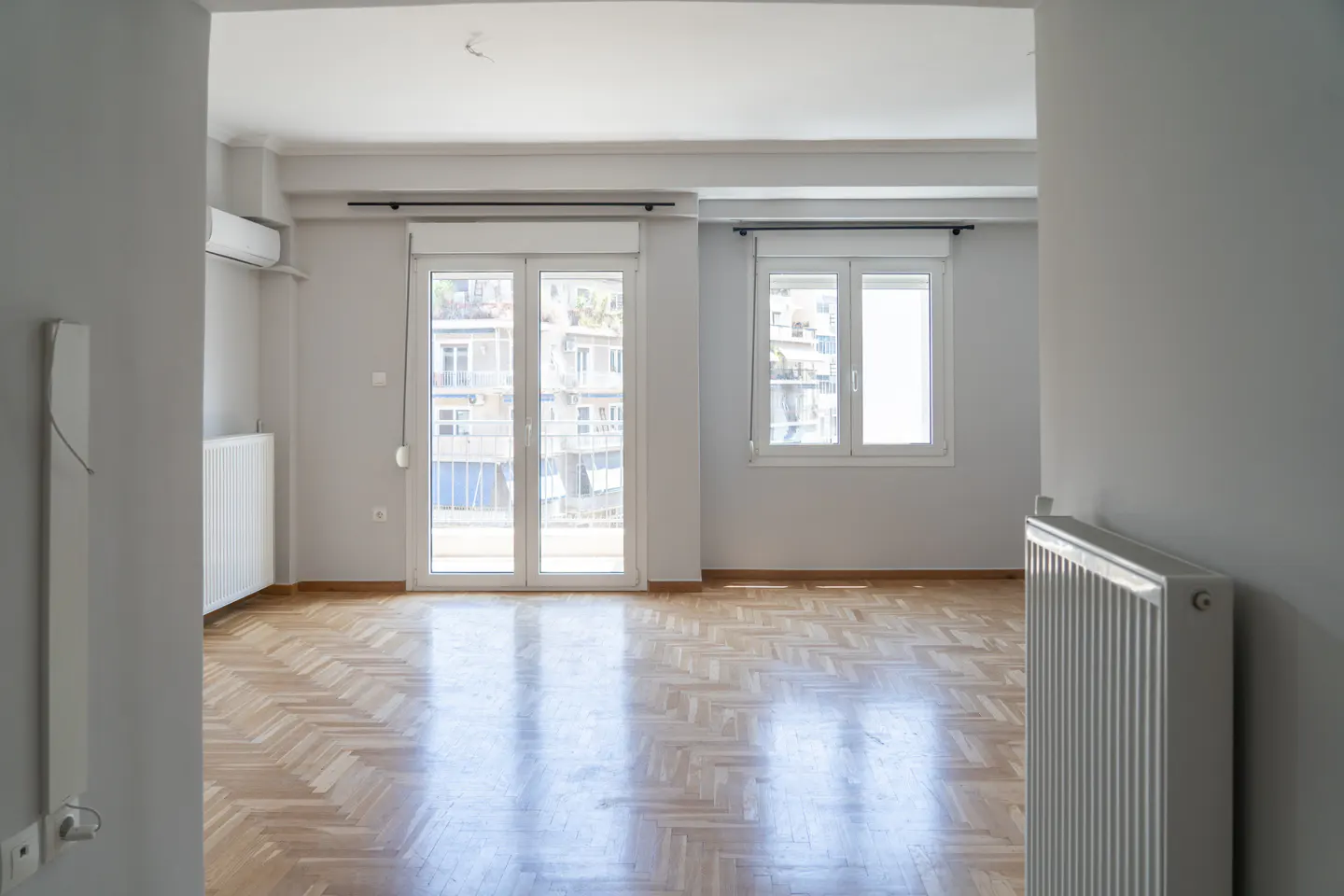 Bright, empty room with herringbone wood floors, white walls, radiator, and balcony doors.