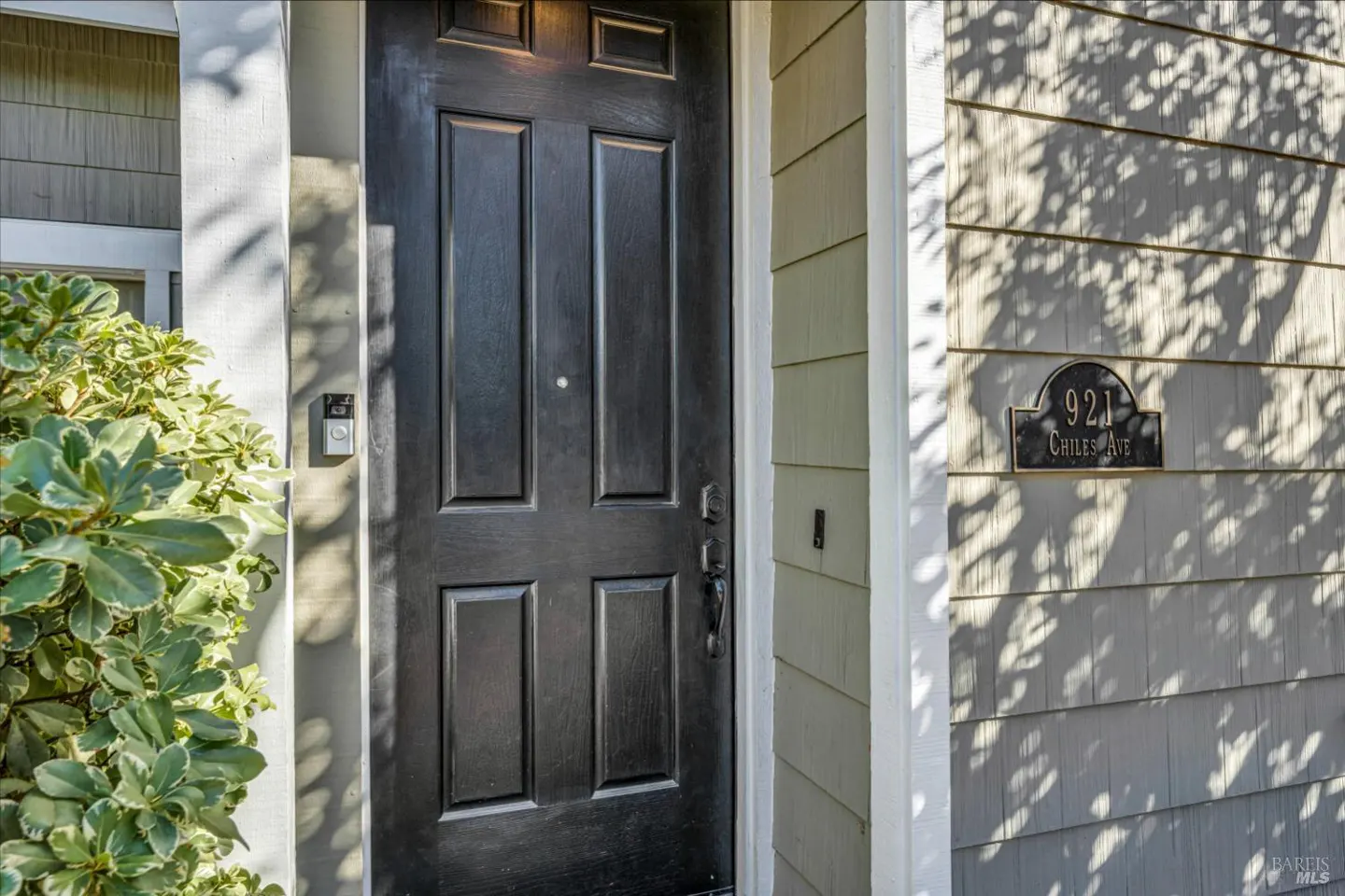 Front door of a house with a black door, light gray siding, and a bush on the left. The address, 921 Chiles Ave, is on the siding.