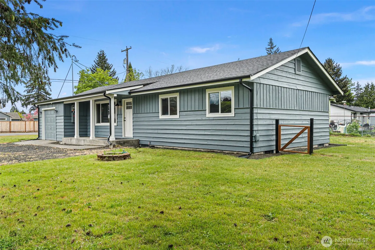 A single-story, light blue house with white trim and a gray roof sits on a green lawn under a blue sky. A small wooden gate is visible on the side.