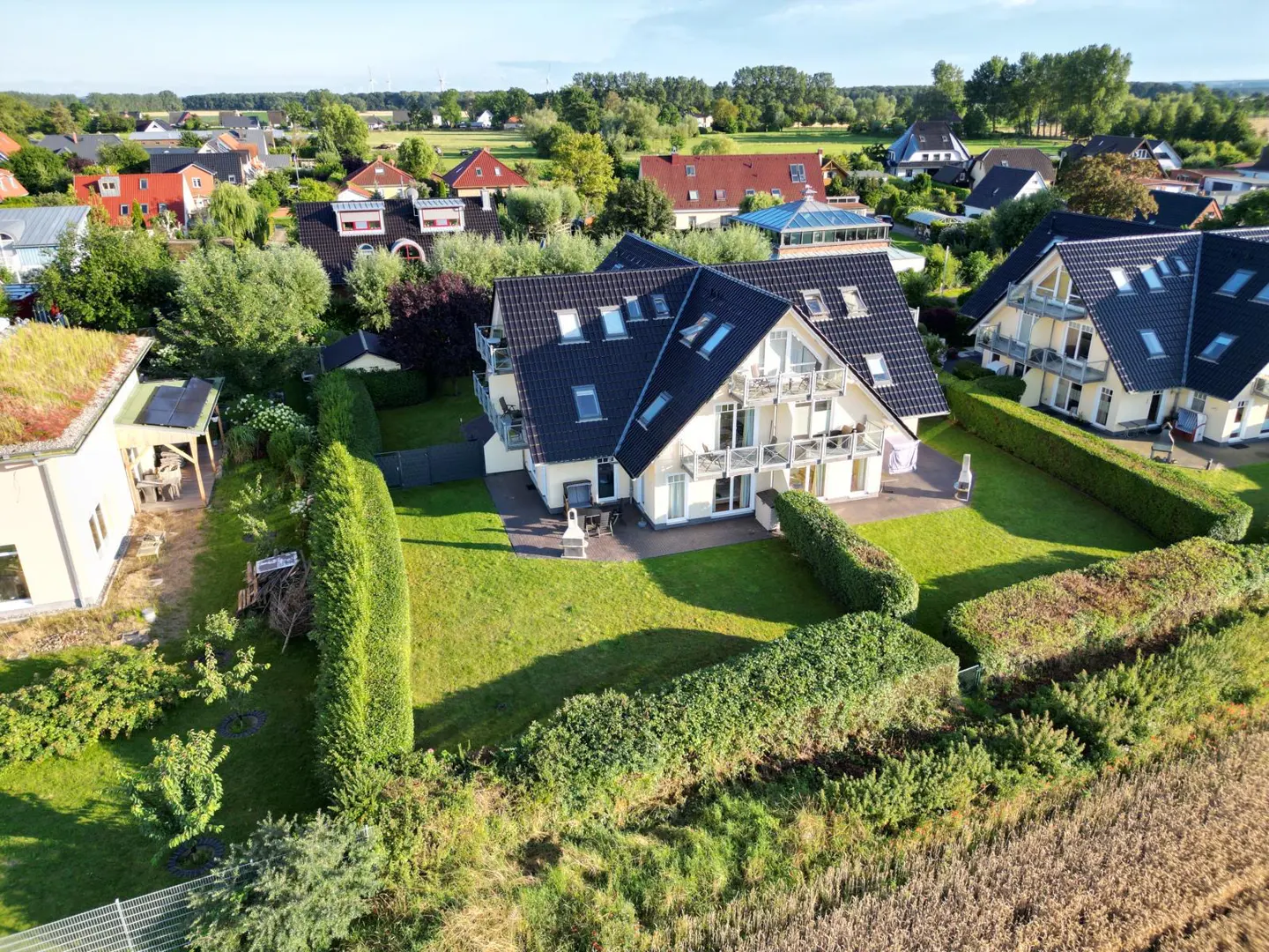 Aerial view of a modern, two-story house with a black roof and white facade, surrounded by green lawns and trimmed hedges in a residential area.