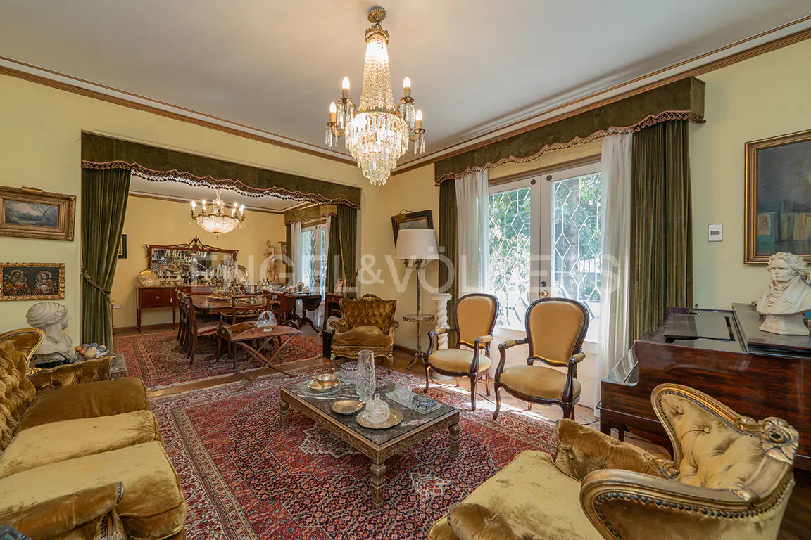 A living room with yellow walls, a crystal chandelier, and antique furniture. A red rug covers the floor.