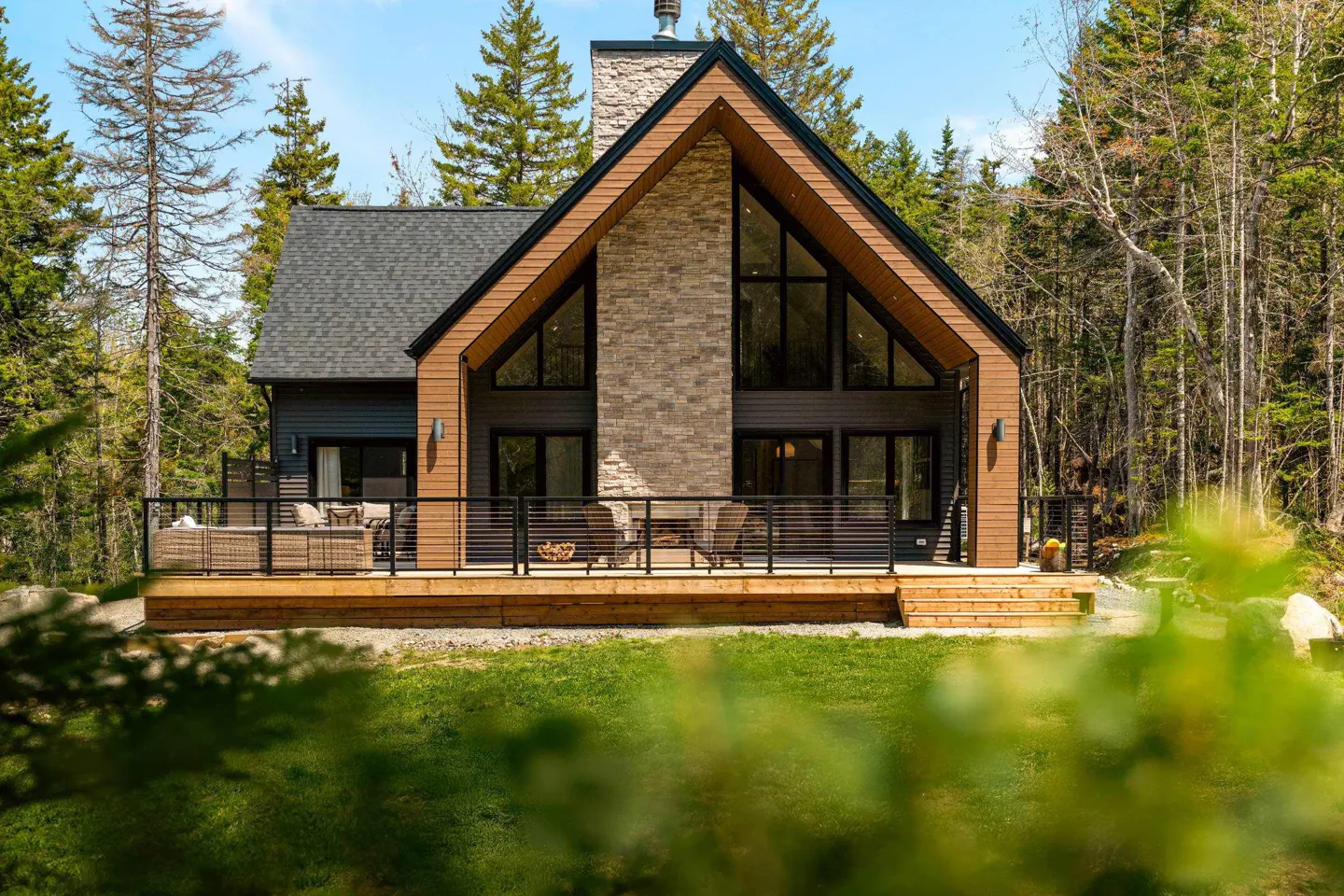 Modern A-frame cabin with a stone chimney, large windows, and a wooden deck, surrounded by a lush forest.