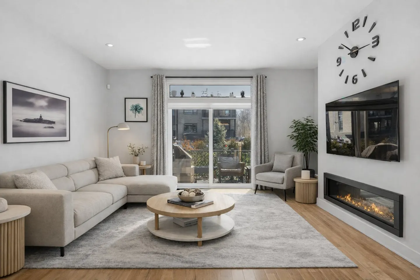 Bright living room with a beige sectional sofa, round wood coffee table, and a modern fireplace under a large TV. A sliding glass door leads to a balcony.